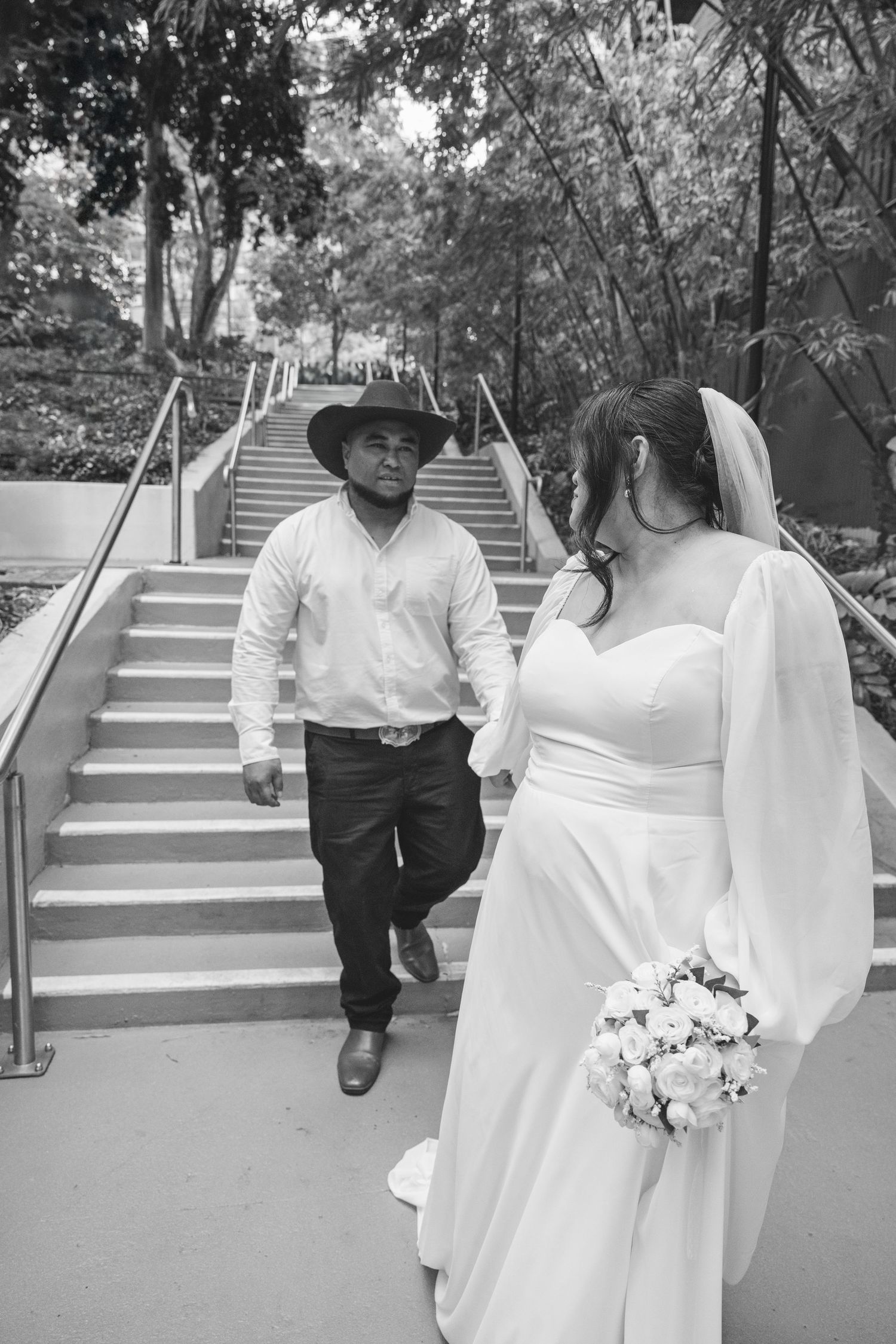 Black and white photo of bride in flowing dress and person in casual attire meeting on outdoor staircase surrounded by trees.