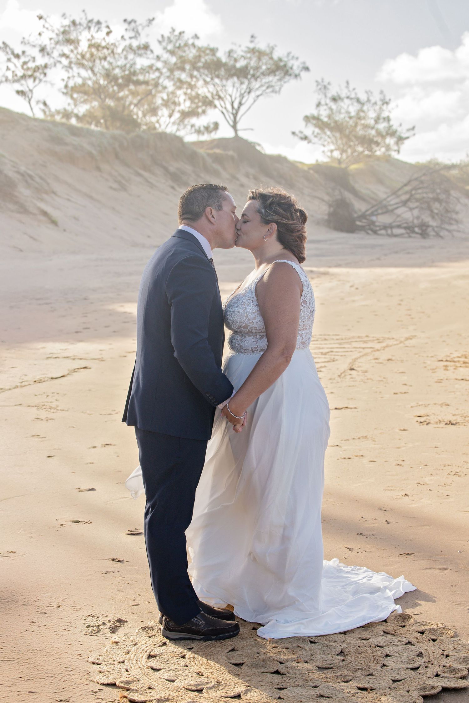 A romantic couple in wedding attire share a tender kiss on a sandy beach with trees in the background.