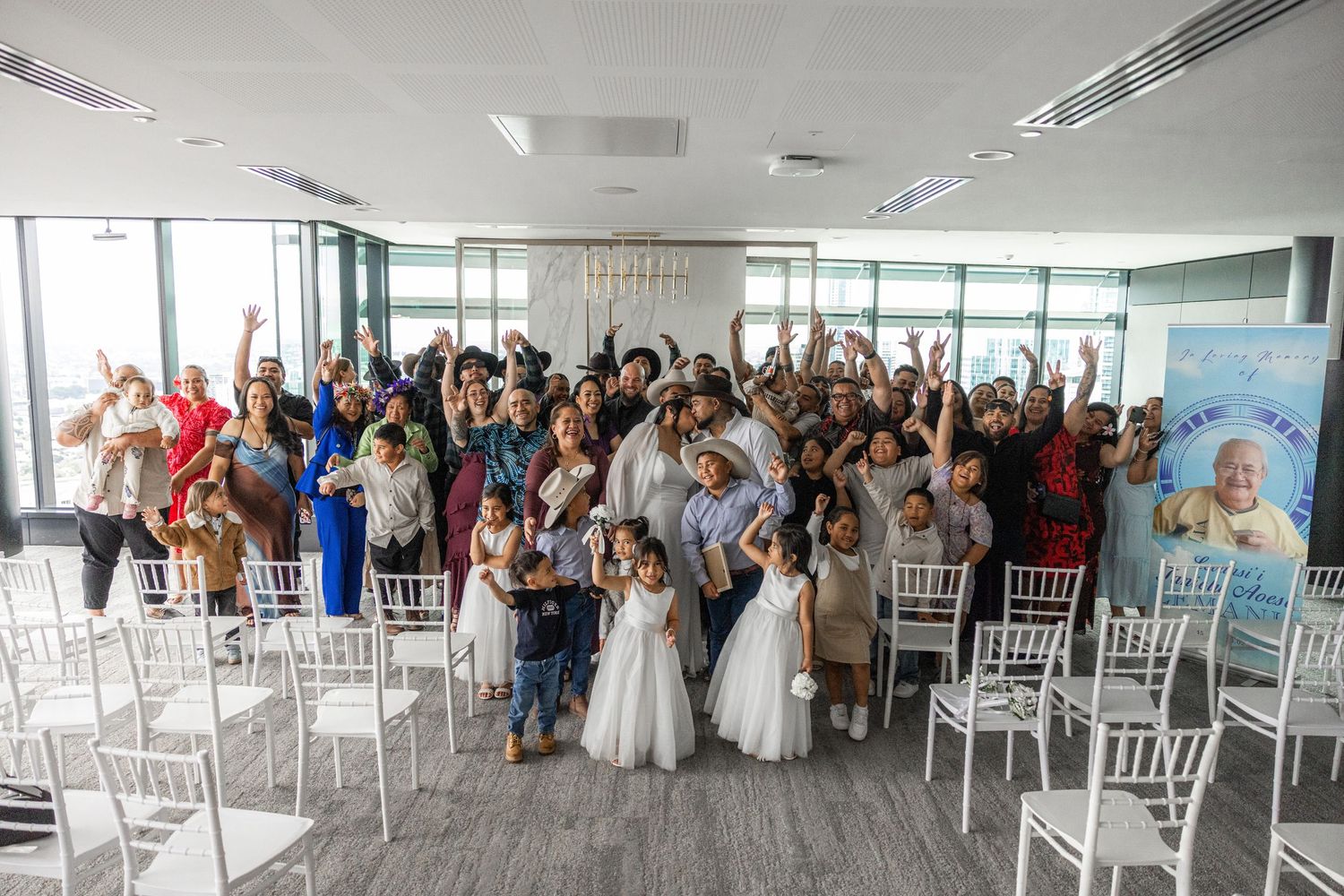 Wedding guests celebrating with raised hands in a bright modern venue with white chairs and large windows.