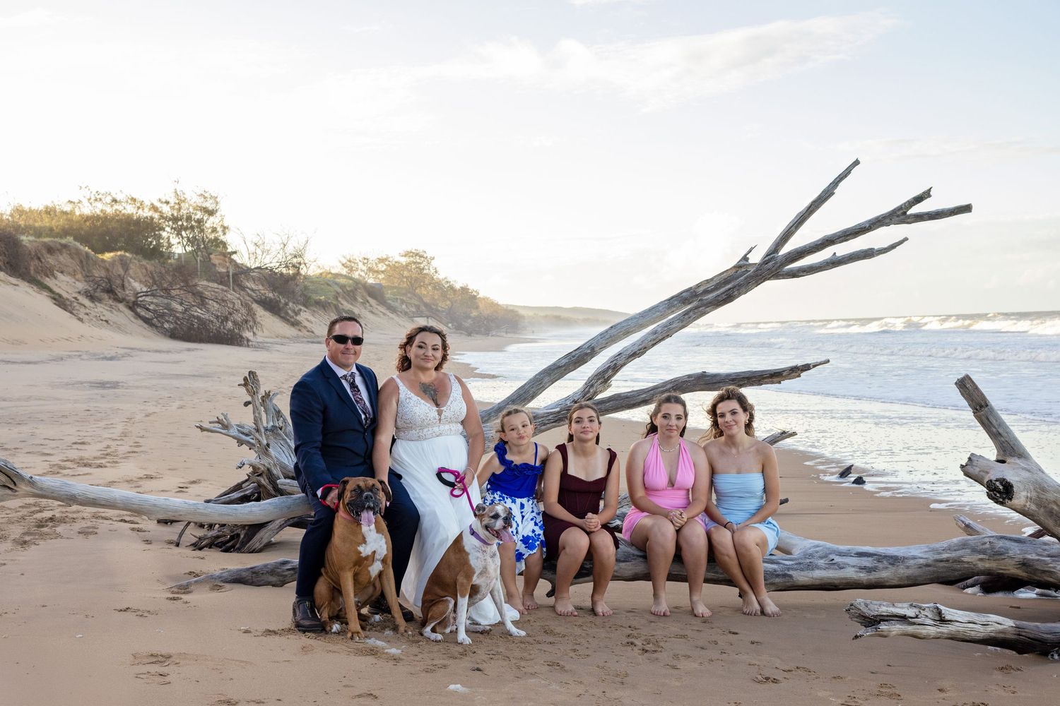 A group poses together on a beach at sunset with driftwood and ocean waves in the background.
