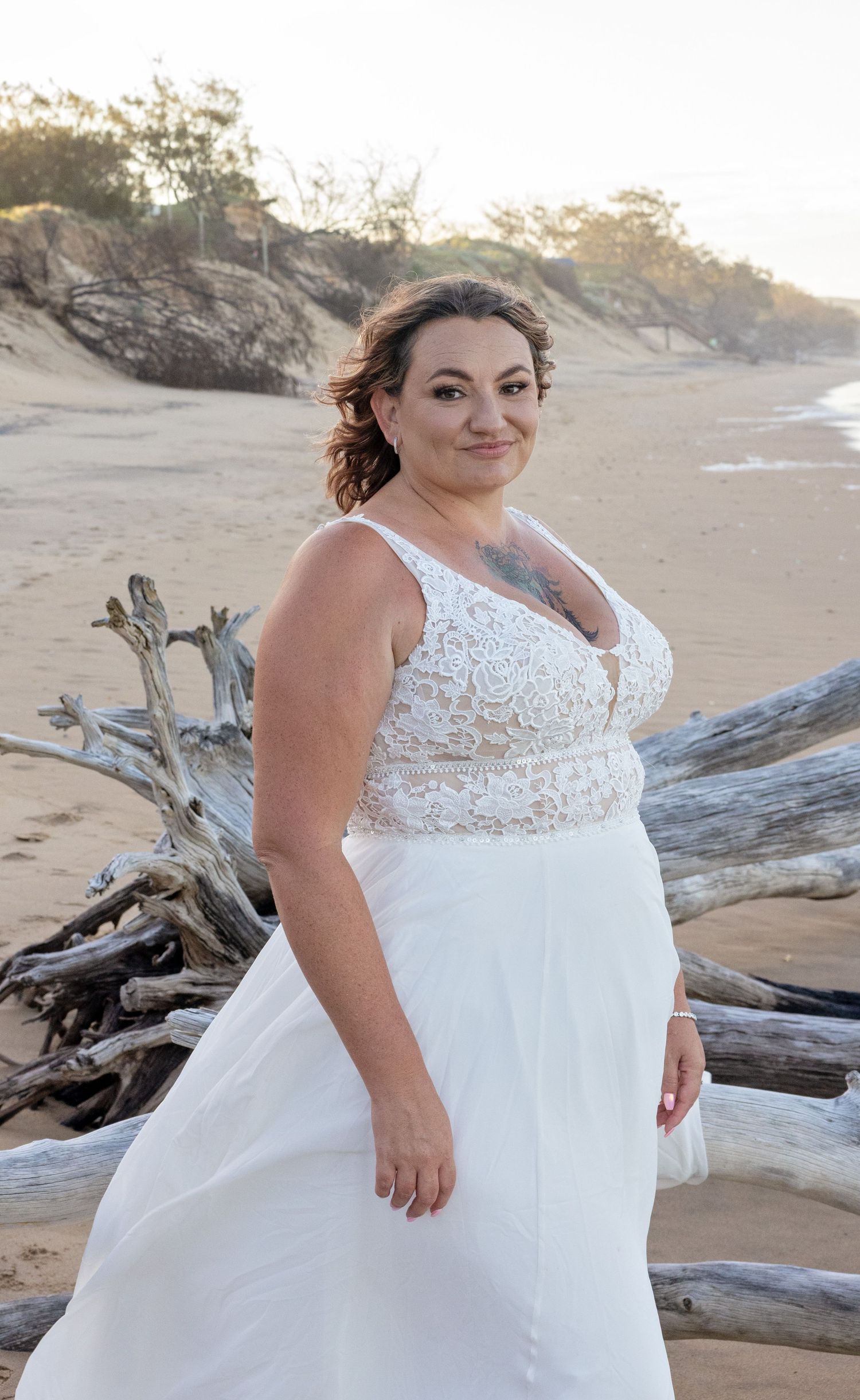 A beautiful wedding dress with lace detail photographed at sunset on a beach with driftwood and sand dunes in background.