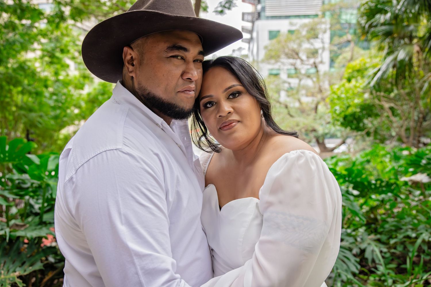 A couple embraces outdoors in a garden setting with the individual wearing a cowboy hat and white clothing.
