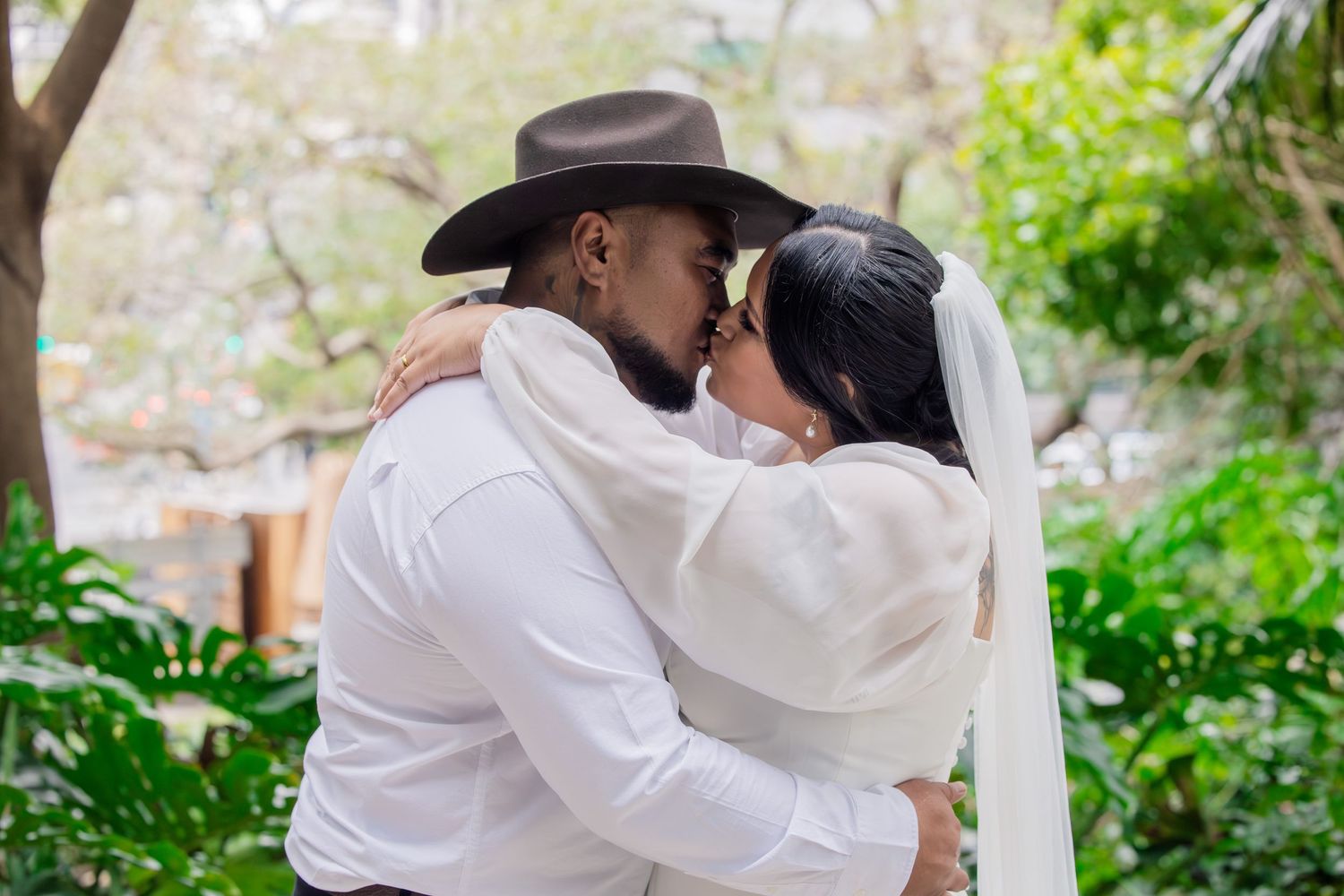 A tender wedding moment captured outdoors with a cowboy hat and flowing white veil against a lush garden backdrop.
