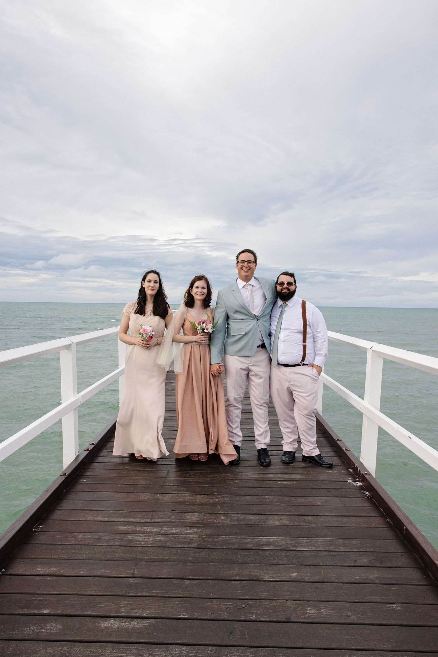 Group in formal attire standing on wooden pier overlooking ocean under cloudy sky.
