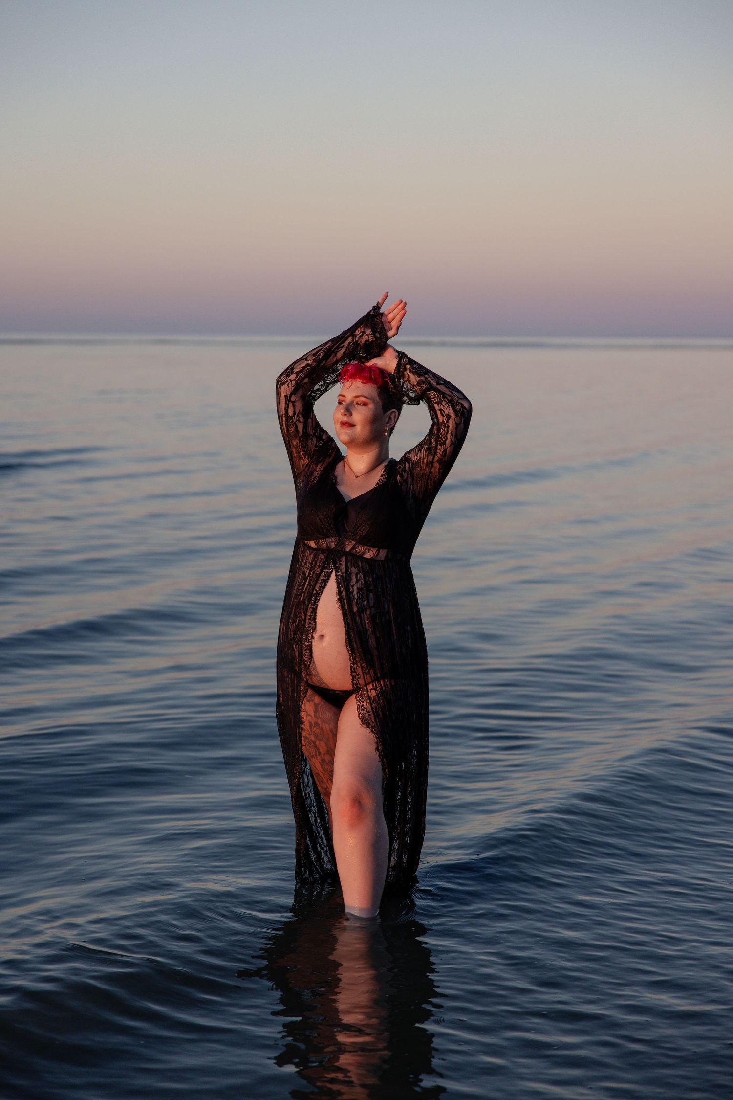 A figure stands in calm ocean waters at sunset wearing black lace against the dusky horizon.