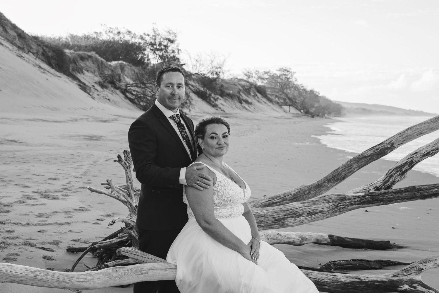A couple in formal wedding attire poses on a beach with driftwood and sand dunes in the black and white scene.