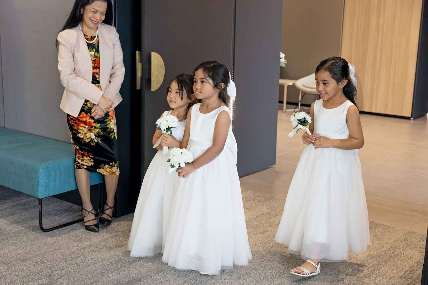 A row of flower girls in white dresses holding bouquets walk across a polished floor during a wedding ceremony.