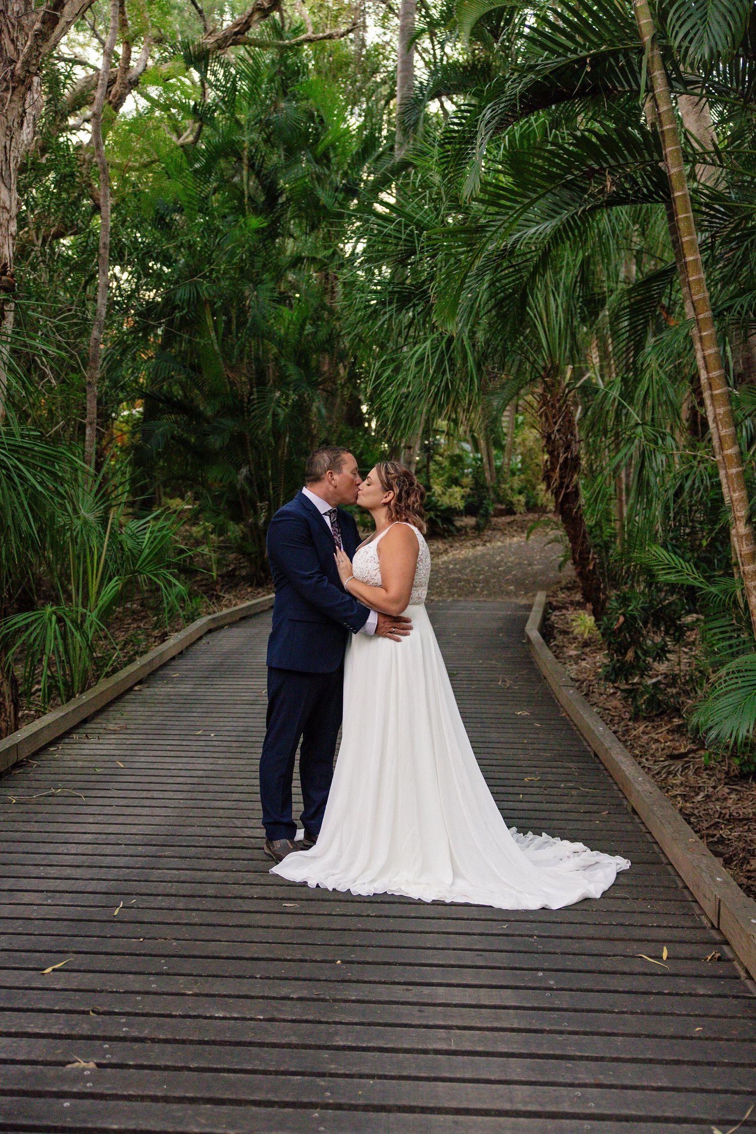 A romantic couple shares a kiss on a wooden boardwalk surrounded by lush tropical palm trees and greenery.