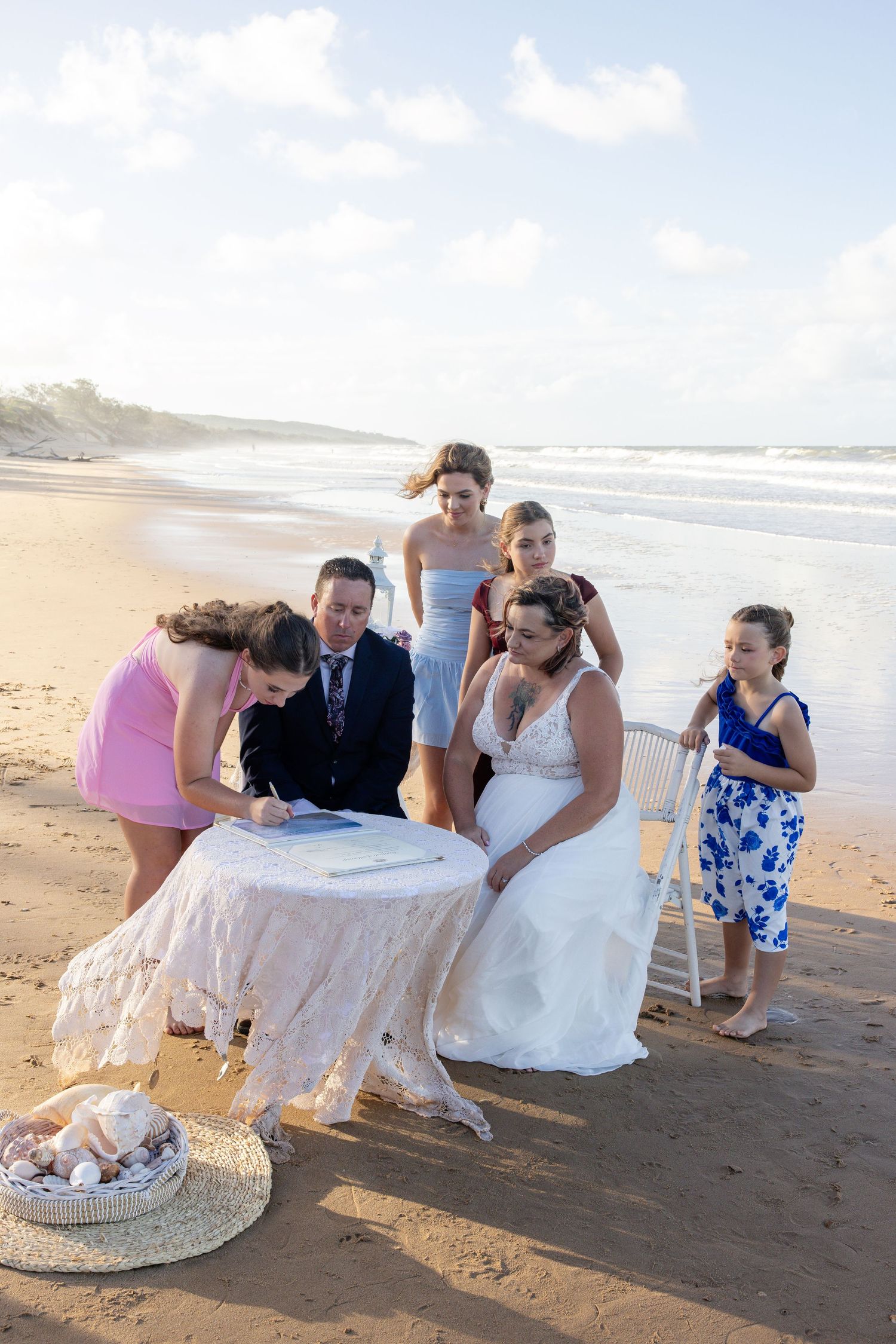 A beach wedding ceremony takes place on the sand with a small group gathered around a table signing documents.