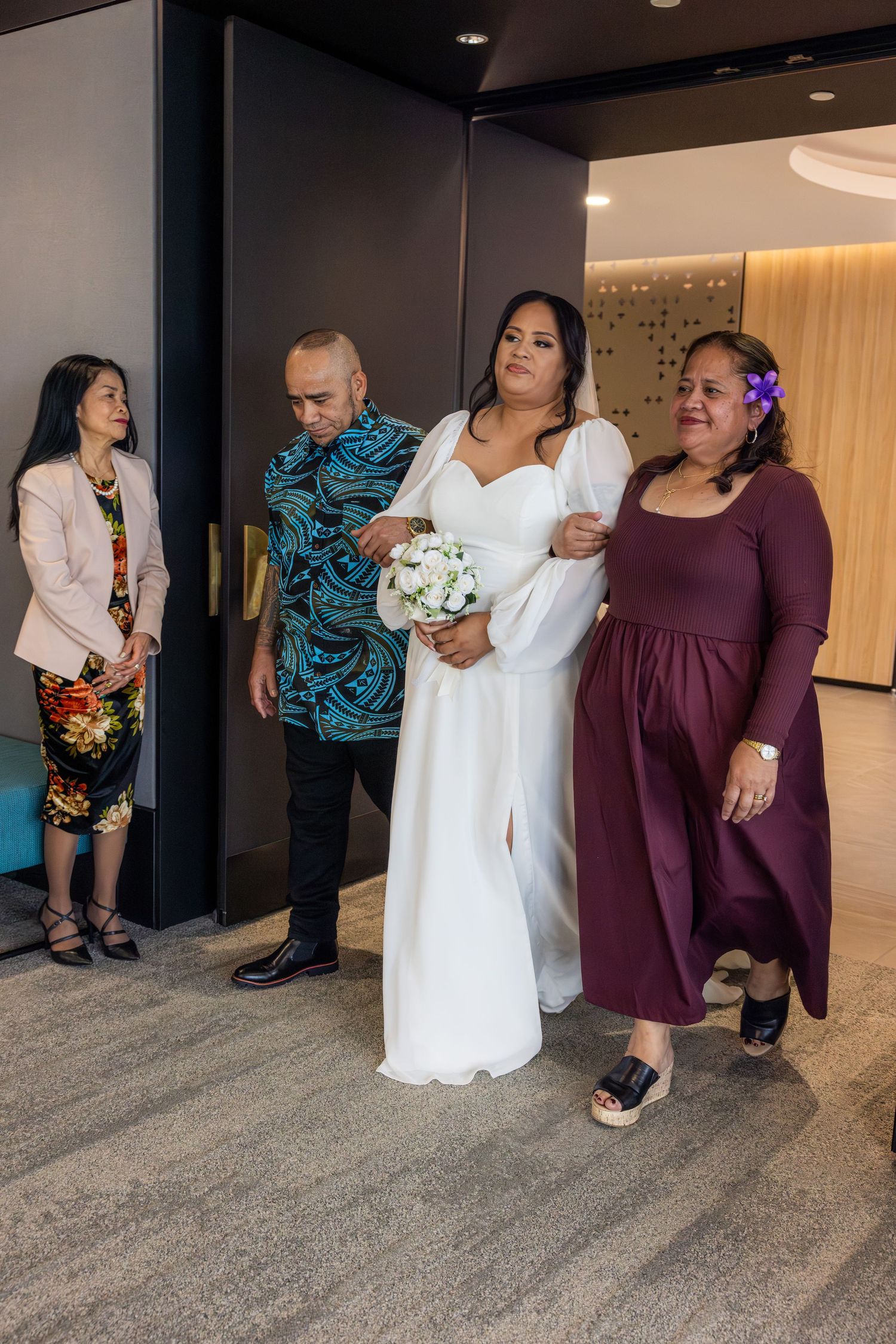 Wedding party members in white and burgundy dresses stand together in a hotel hallway during photo session.
