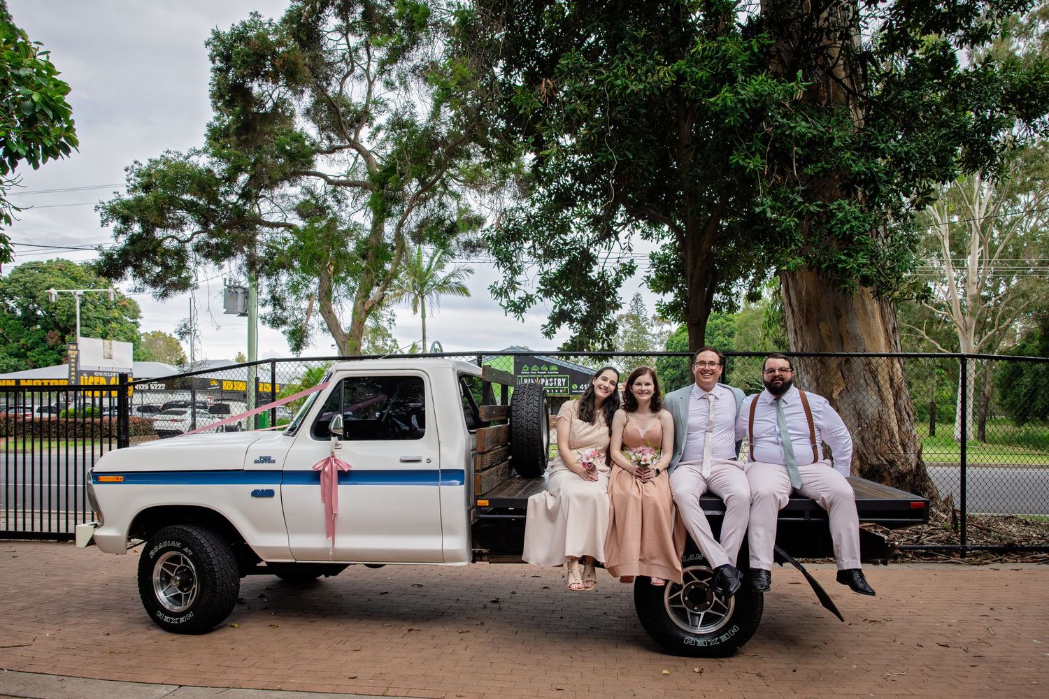 Wedding party poses with vintage white Jeep under large trees in outdoor setting.