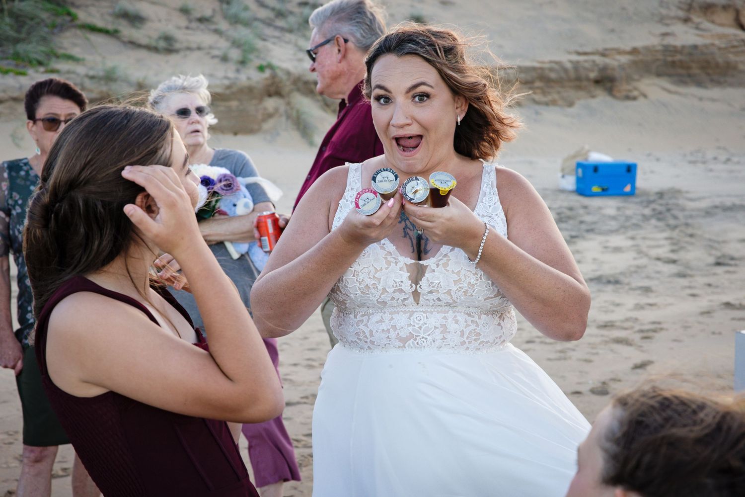 A joyful bride in a white lace dress shares an excited moment with wedding guests during an outdoor beach celebration.