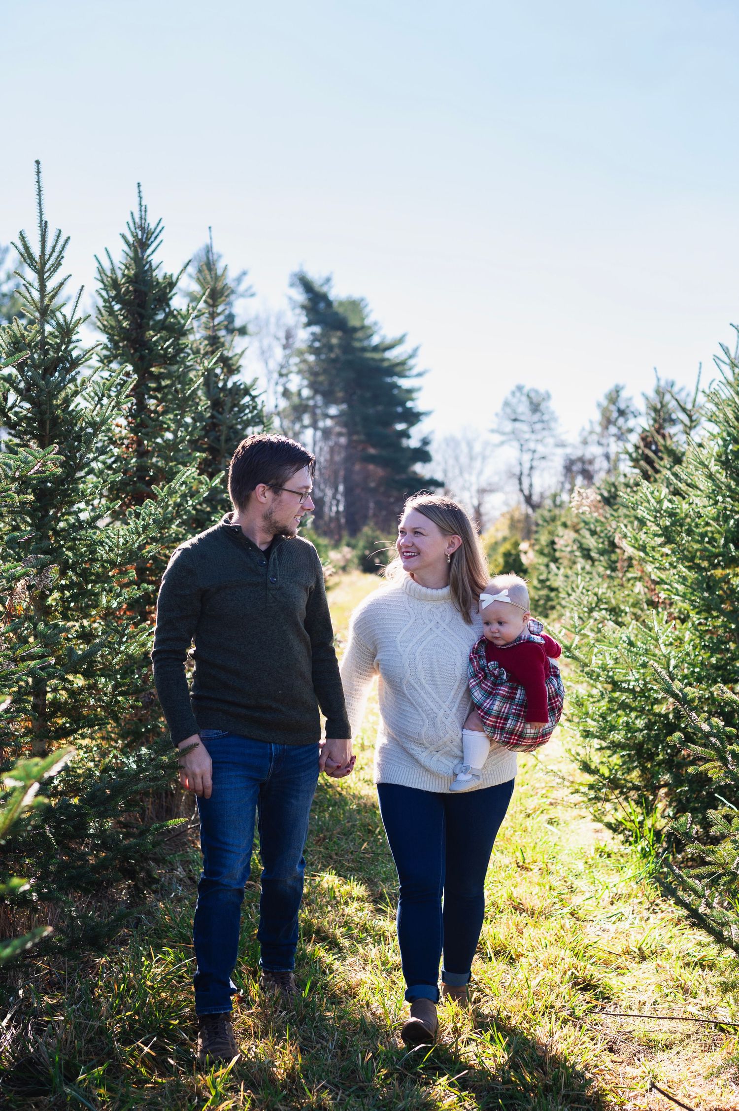Parents walk hand in hand through a Christmas tree farm, mom holding a bundled baby in festive plaid.
