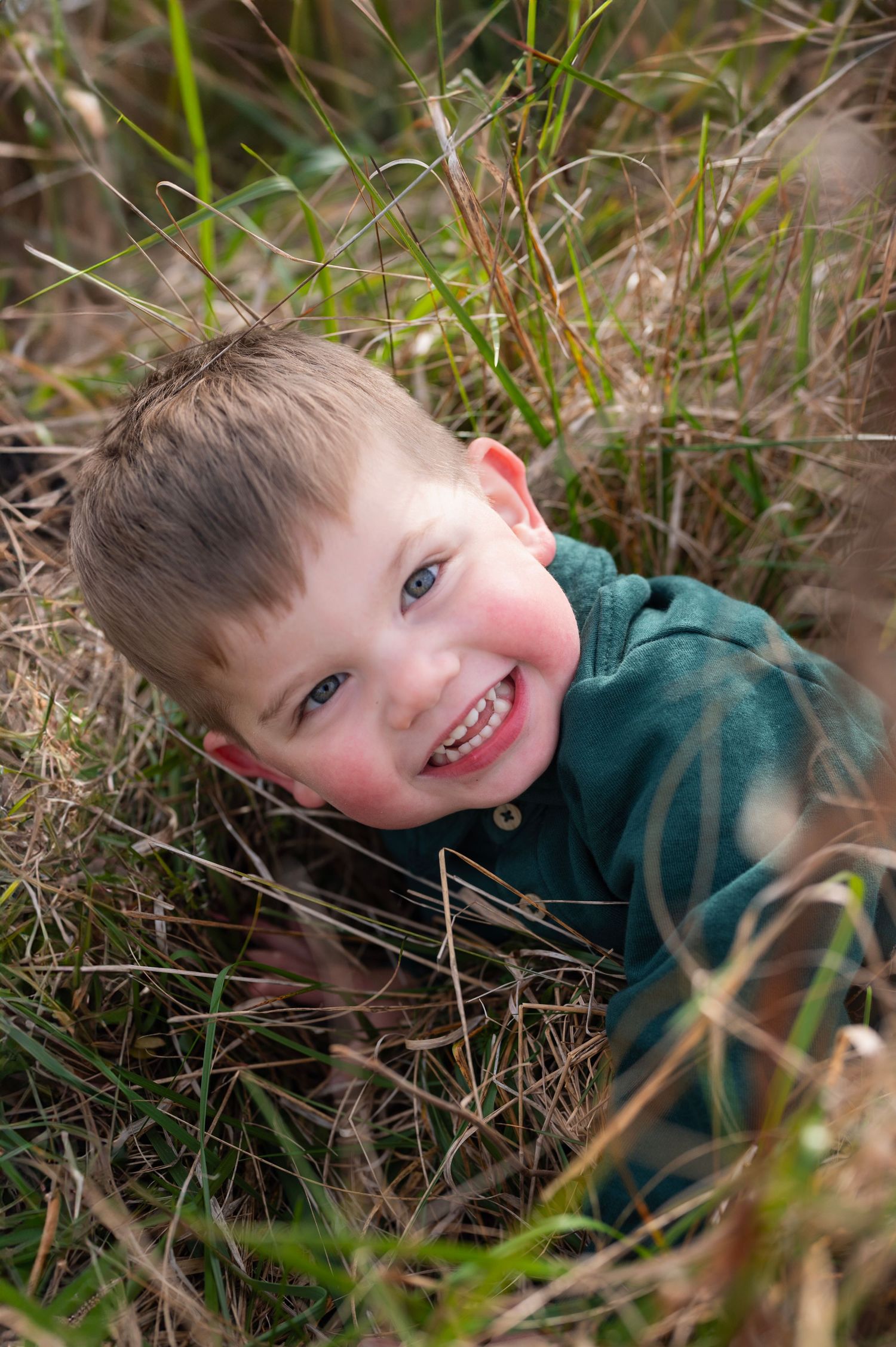 Joyful toddler boy lying in tall grass, looking up with a big smile and rosy cheeks.