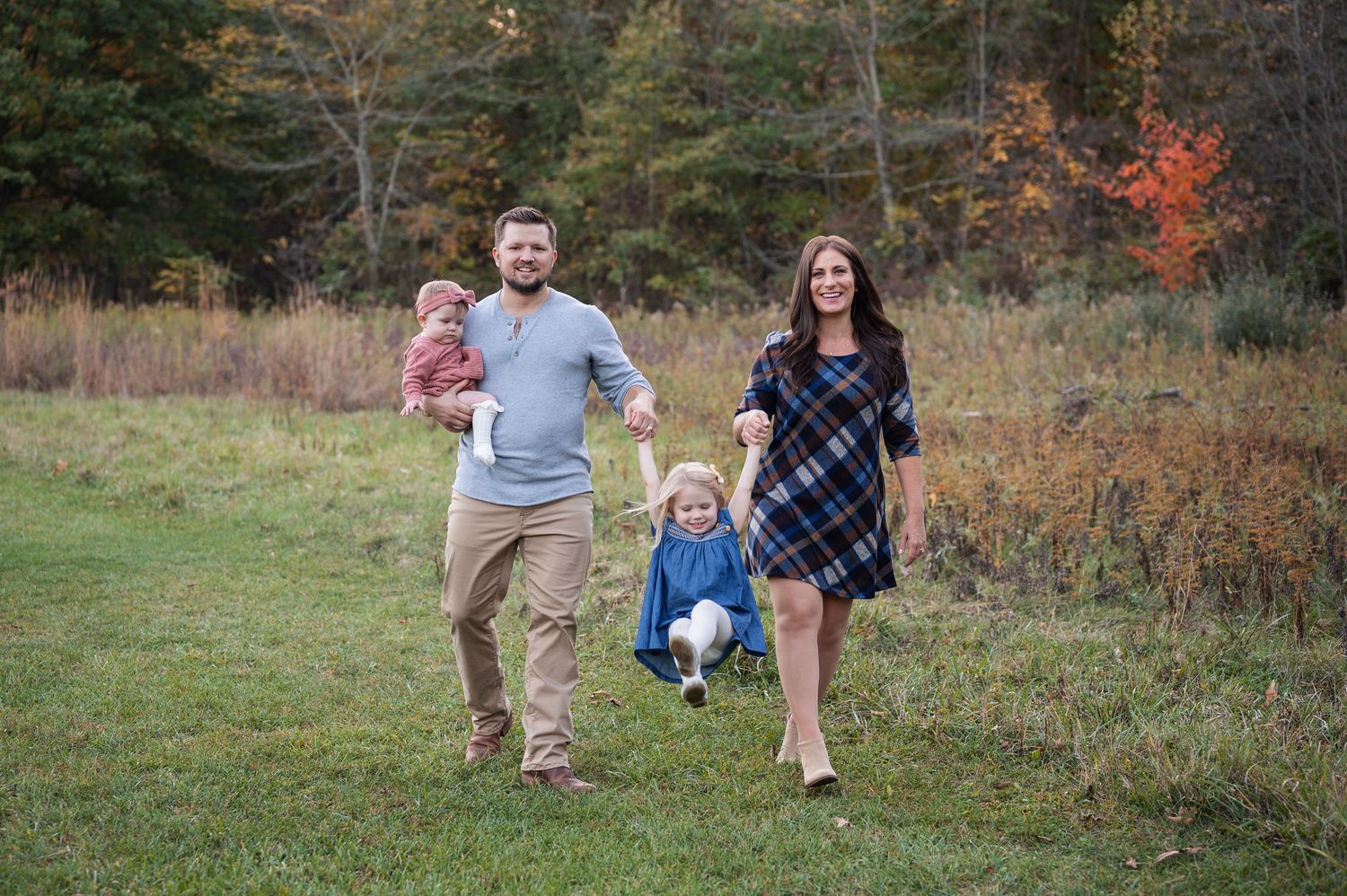 A smiling family walks through a field in fall, lifting their daughter between them while holding their baby.