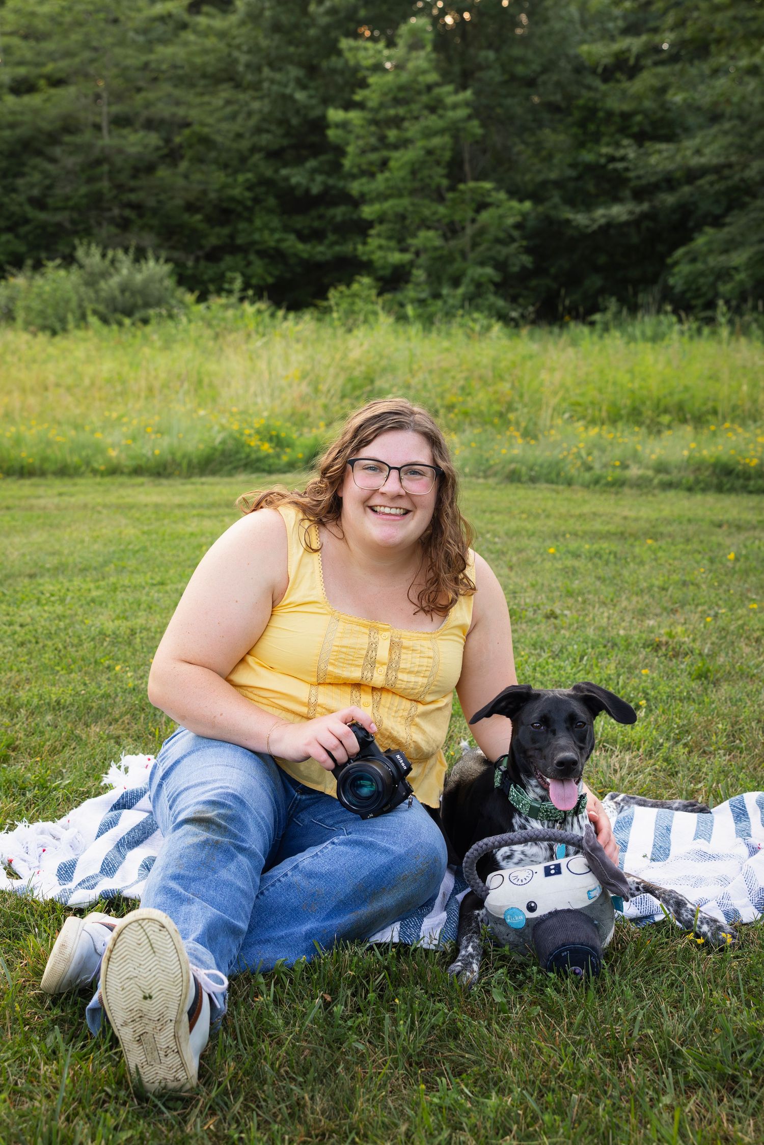 Smiling female photographer in a yellow top sitting on a blanket in a field with her playful black dog and a camera-themed plush toy.