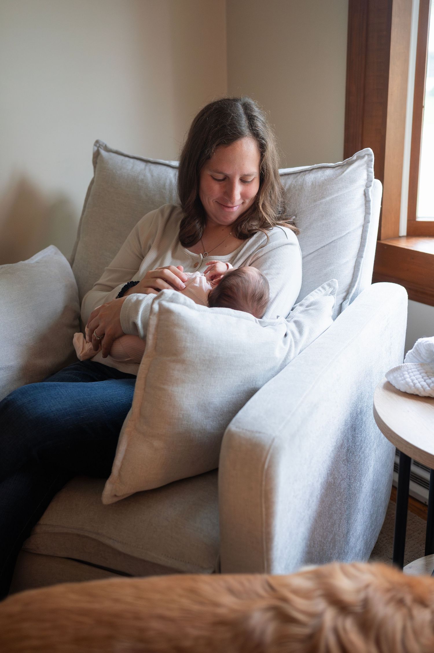 Mother lovingly bottle-feeding her newborn in a cozy chair near a window during an in-home session.