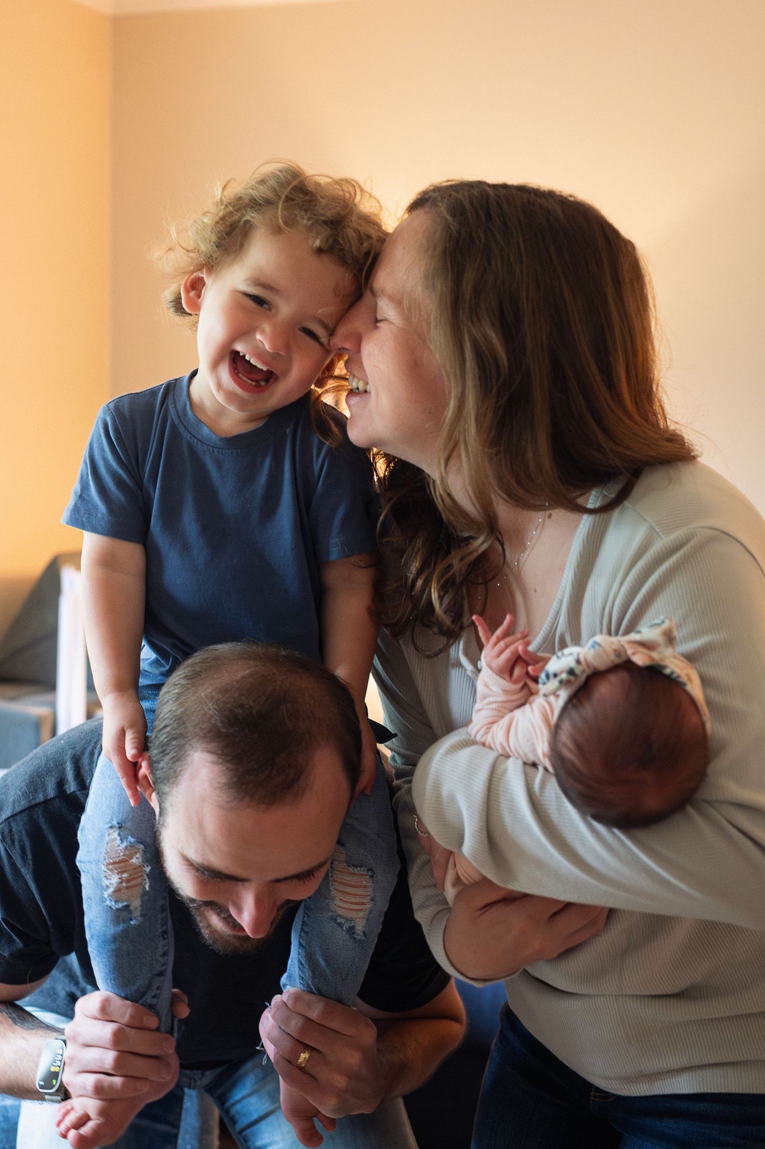 Joyful moment with toddler laughing on dad’s shoulders and mom cradling newborn. Real family life photography.