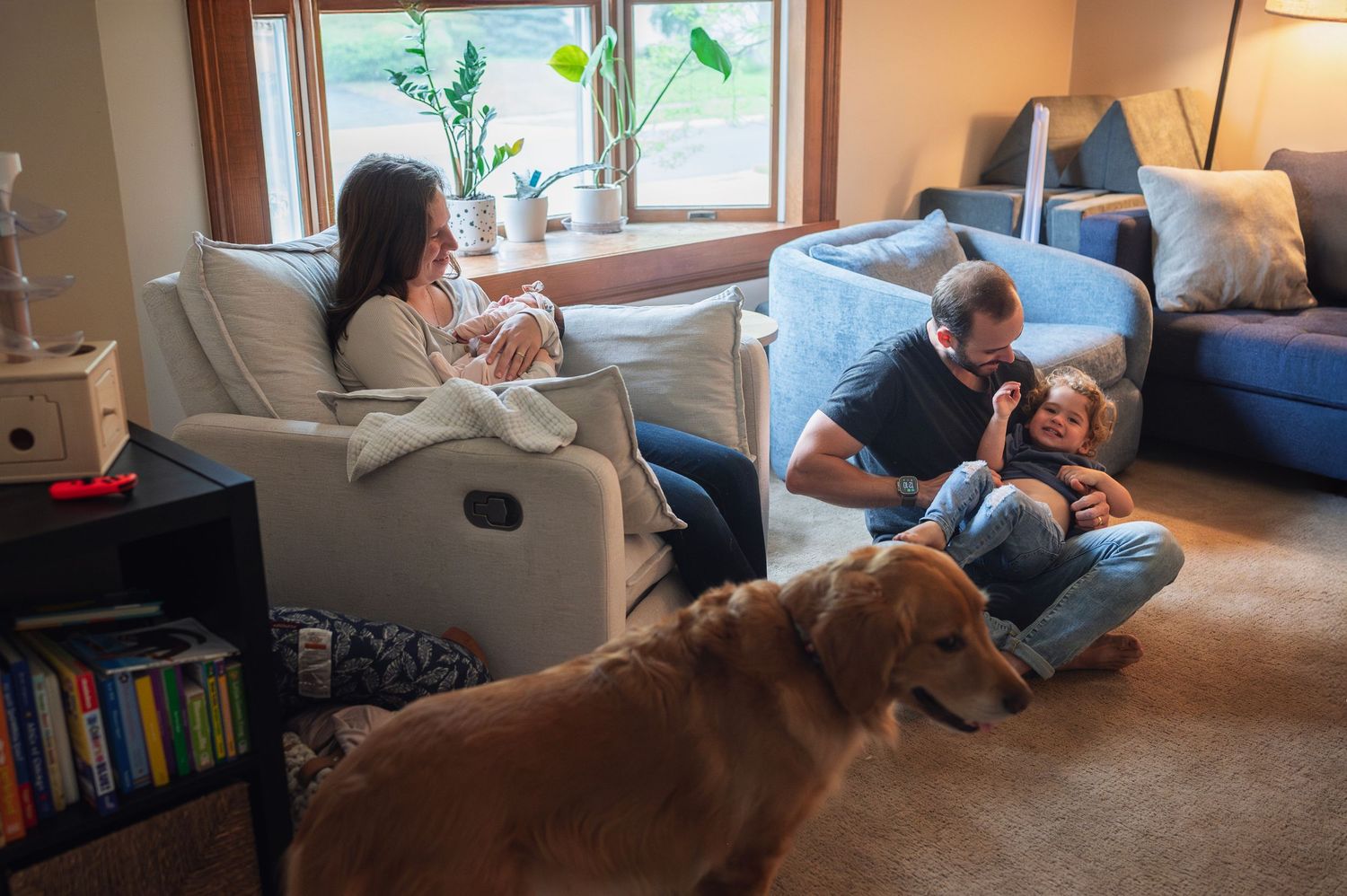 Mom cradles newborn while dad tickles laughing toddler on living room floor, golden retriever watching nearby.