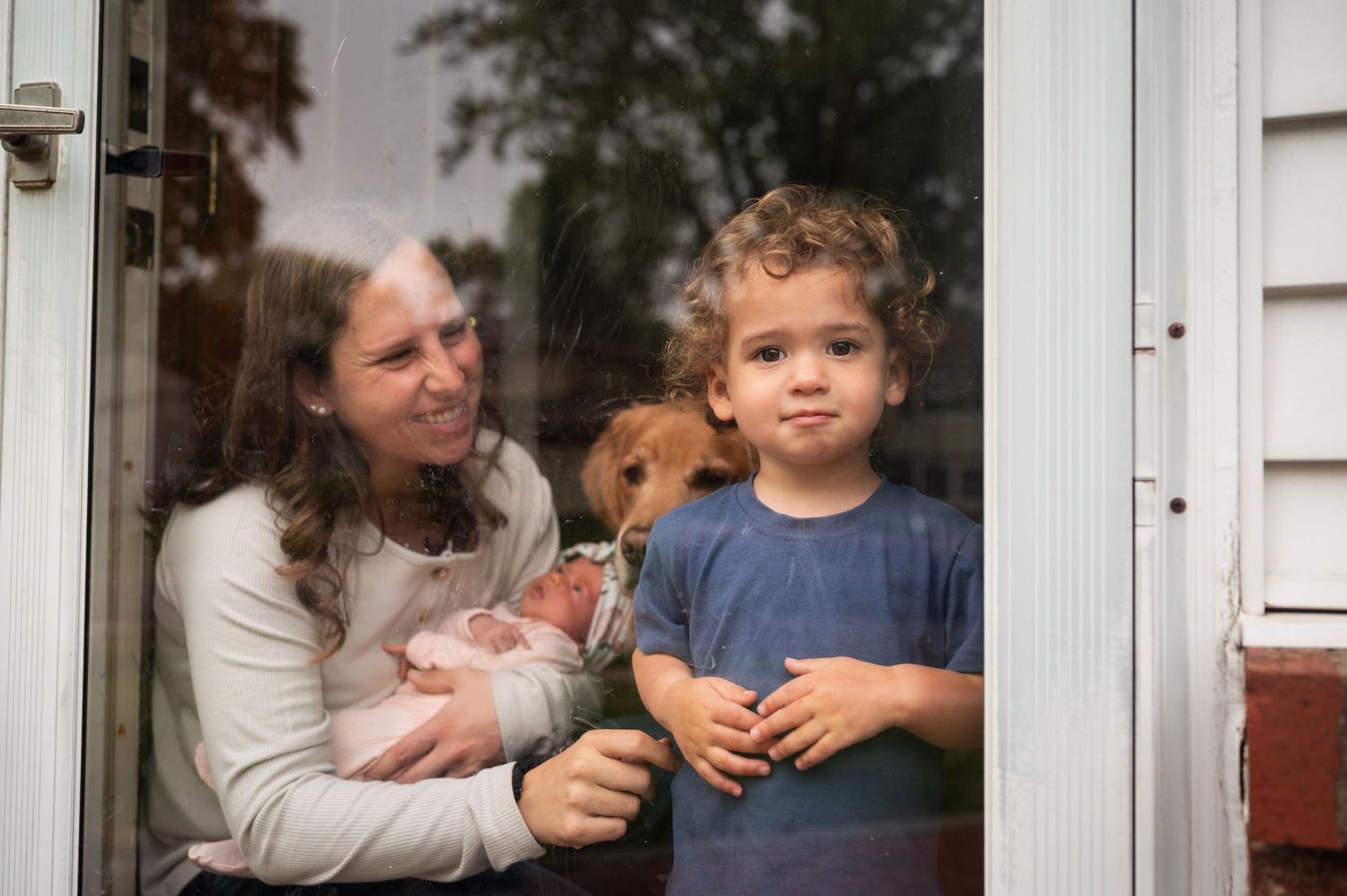 Toddler stands at storm door with serious expression while mom smiles, holding baby and dog peeking through behind.
