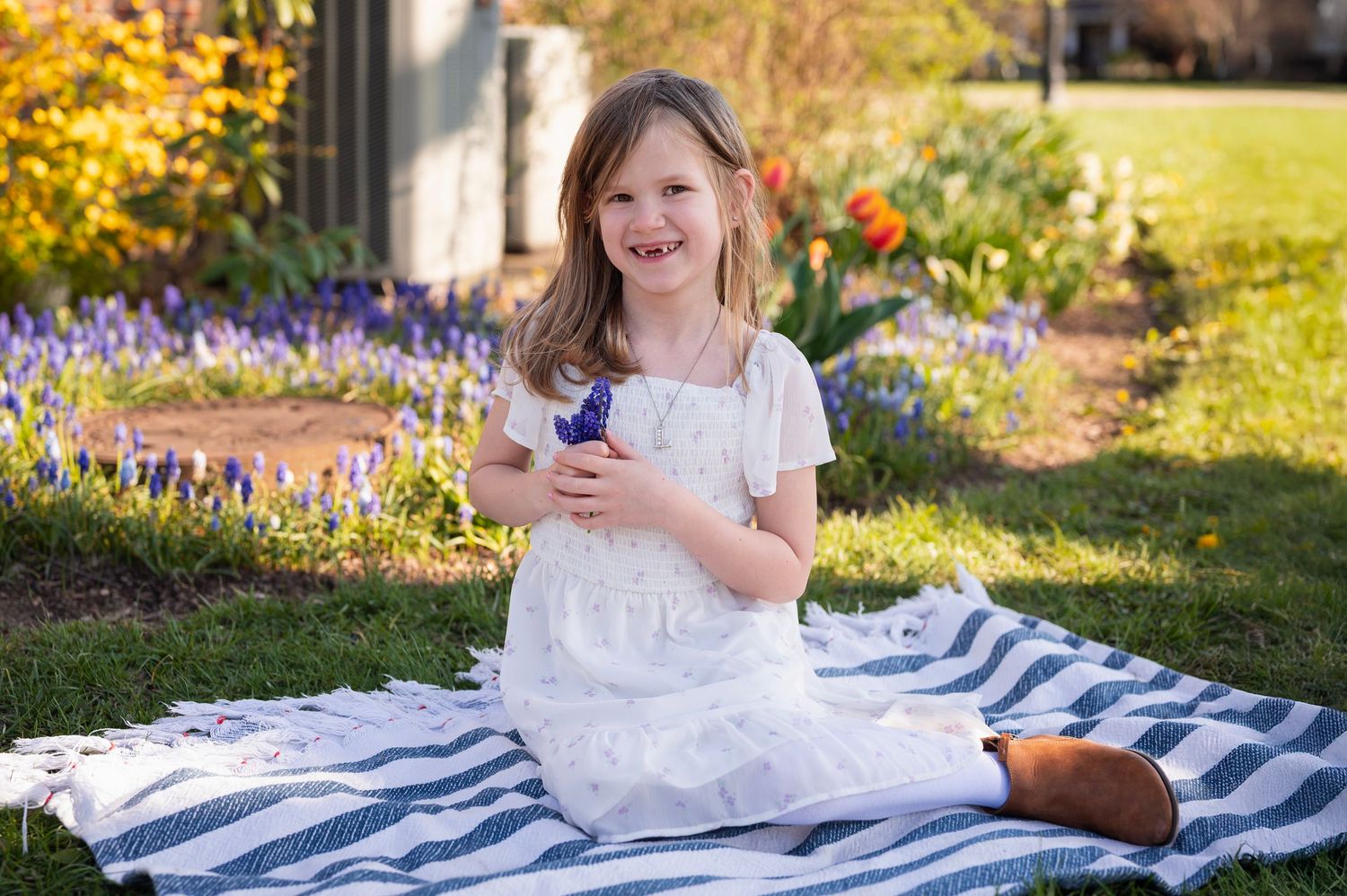 Smiling girl in a white dress holding purple flowers, sitting on a striped blanket in a spring garden.