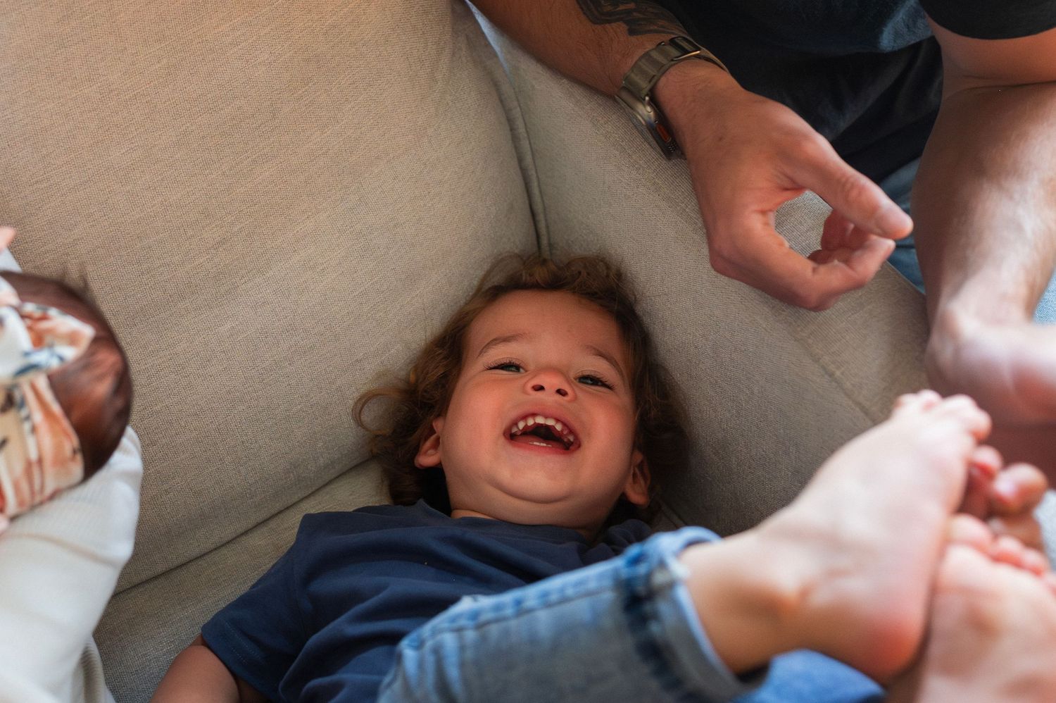 Toddler giggles while lying on couch during lifestyle newborn session with parents nearby.
