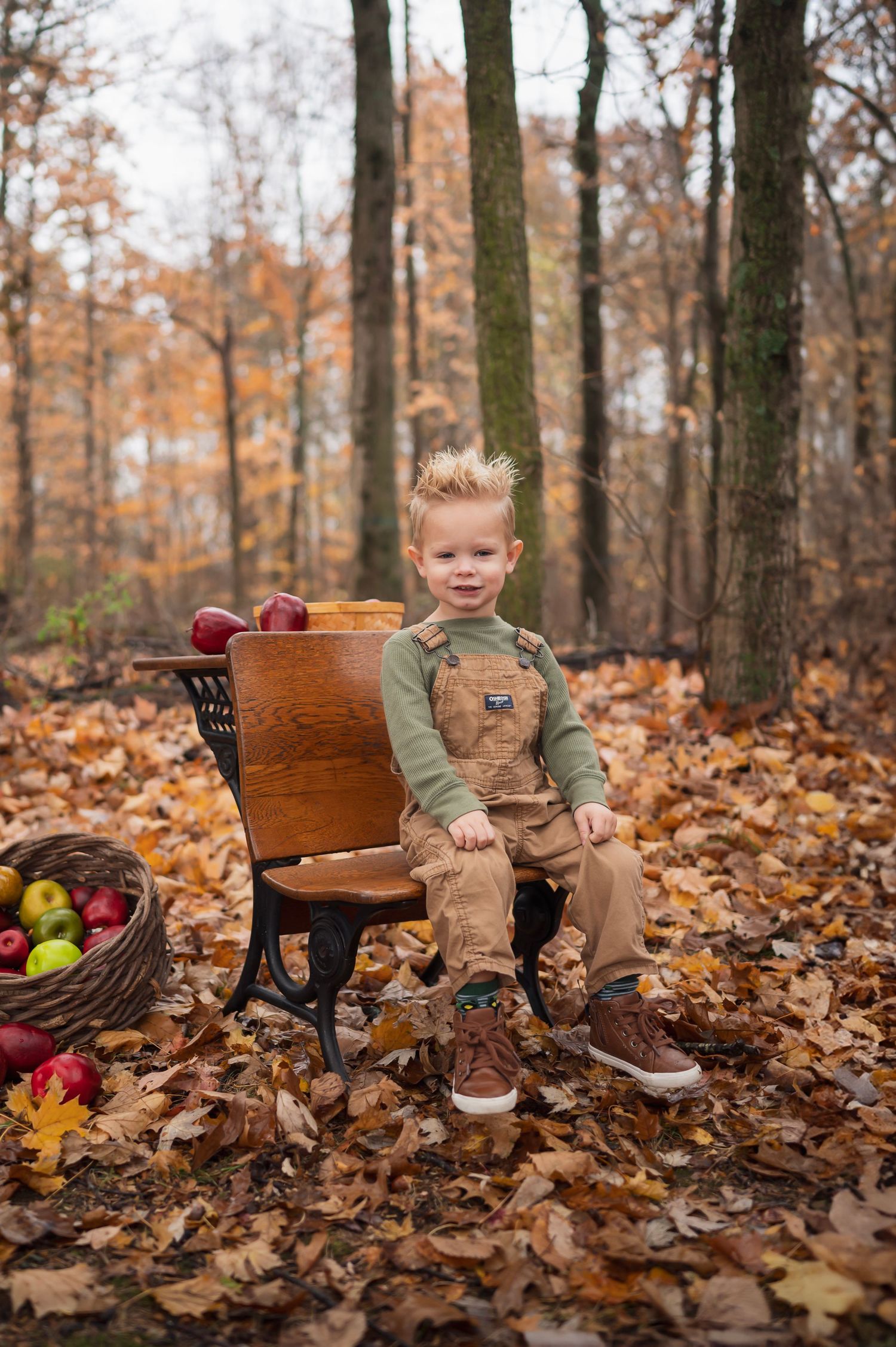 Young boy in overalls sits on a vintage school bench surrounded by autumn leaves and apples during a fall mini session