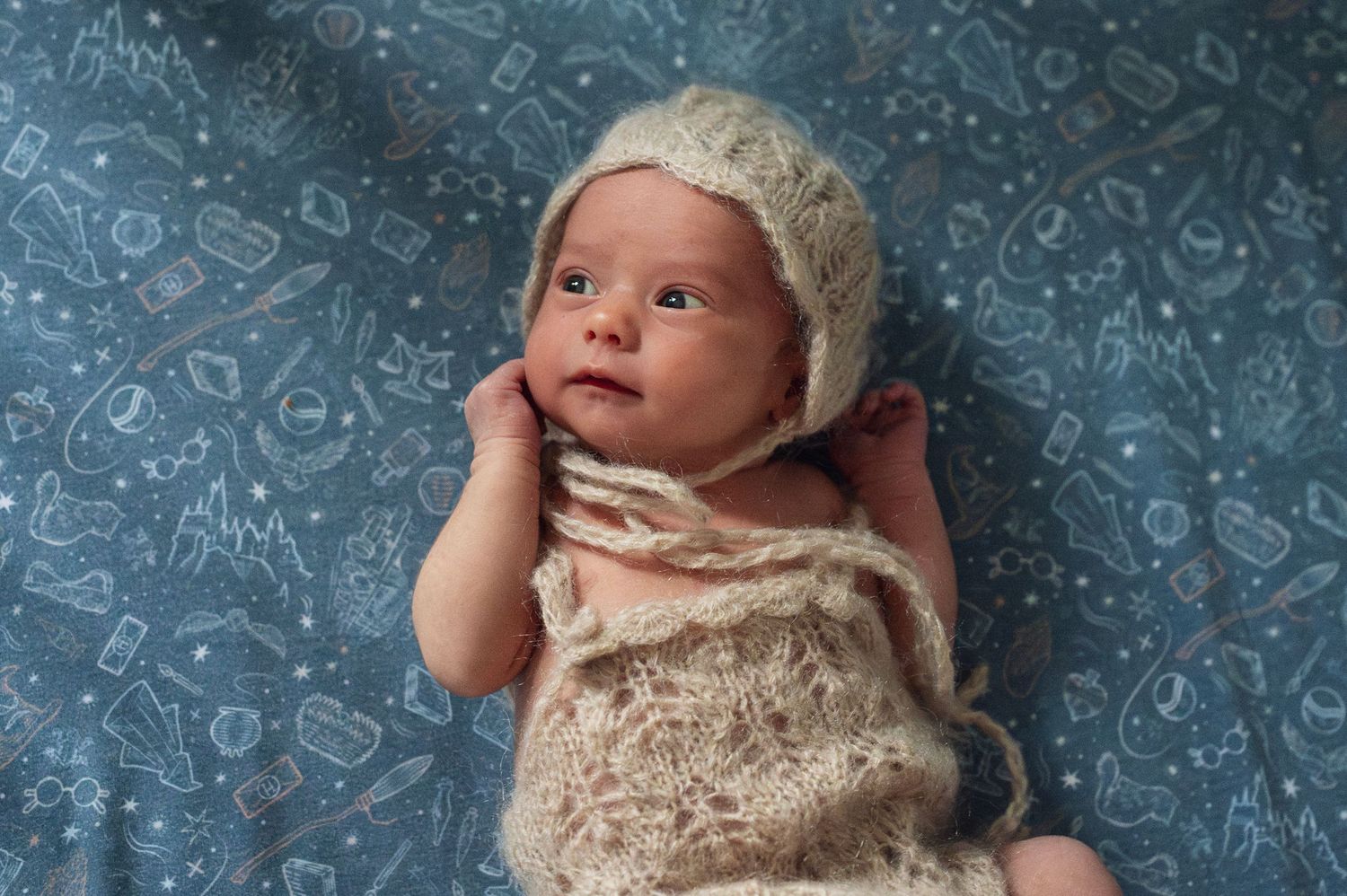 Awake newborn in a knitted bonnet lying on a whimsical, illustrated blanket during a home photo session.