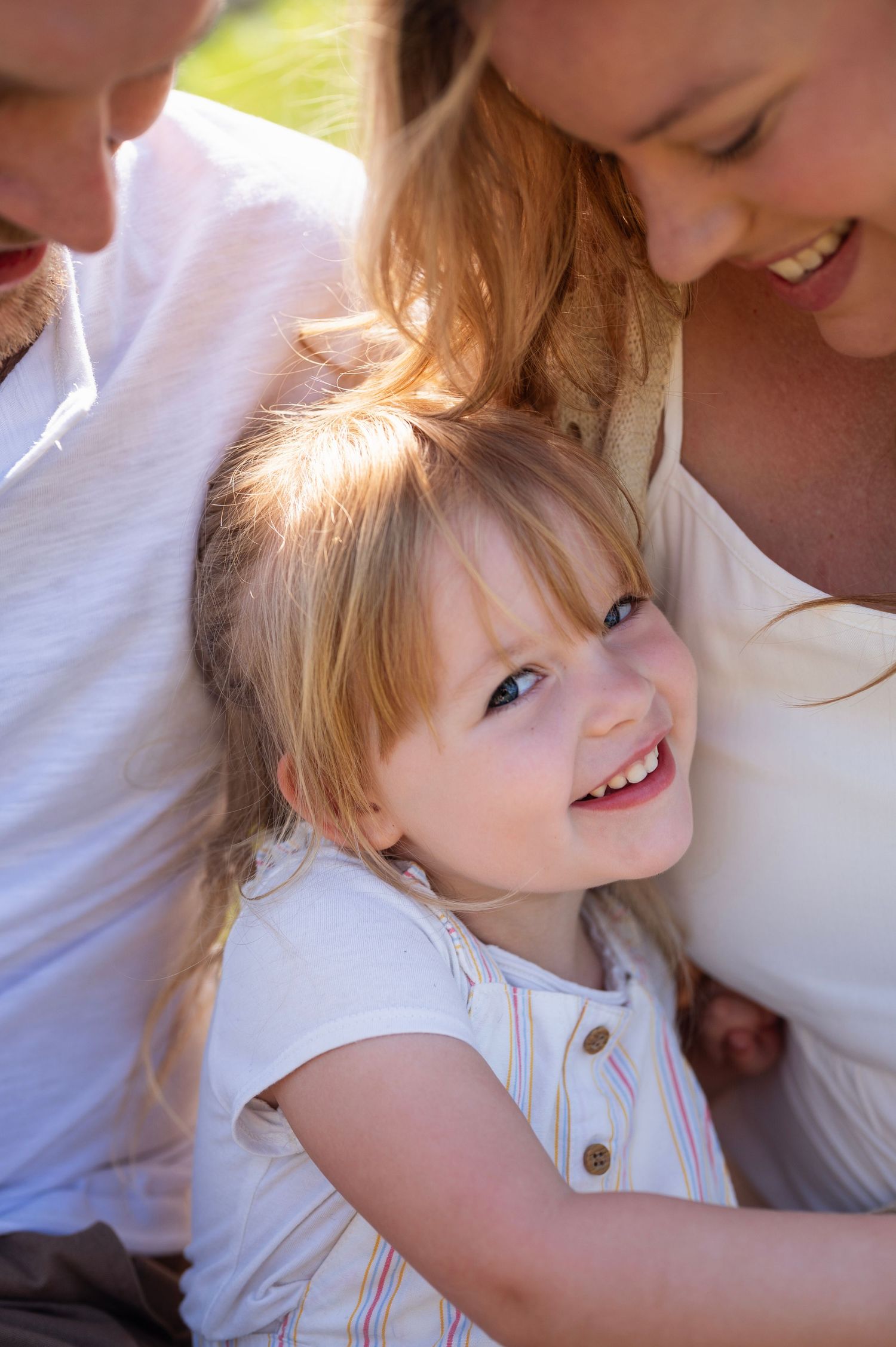 Young girl nestled between her parents, grinning up at the camera with sun-kissed hair and soft light.
