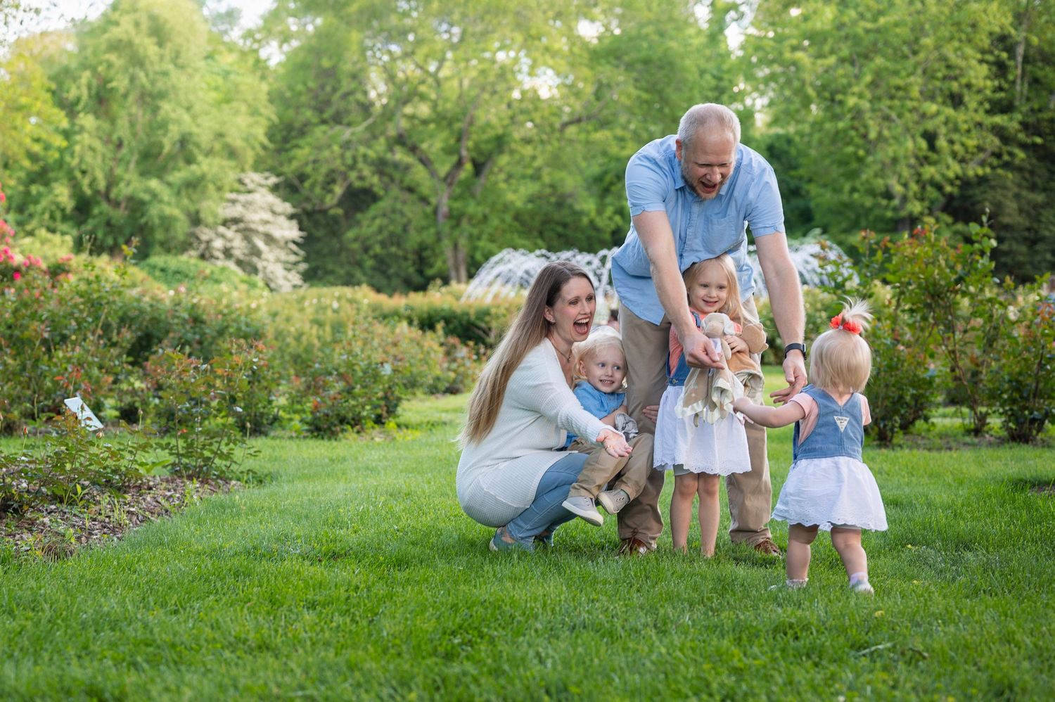 Family encourages toddler walking toward them during a spring mini session at Park of Roses in Columbus