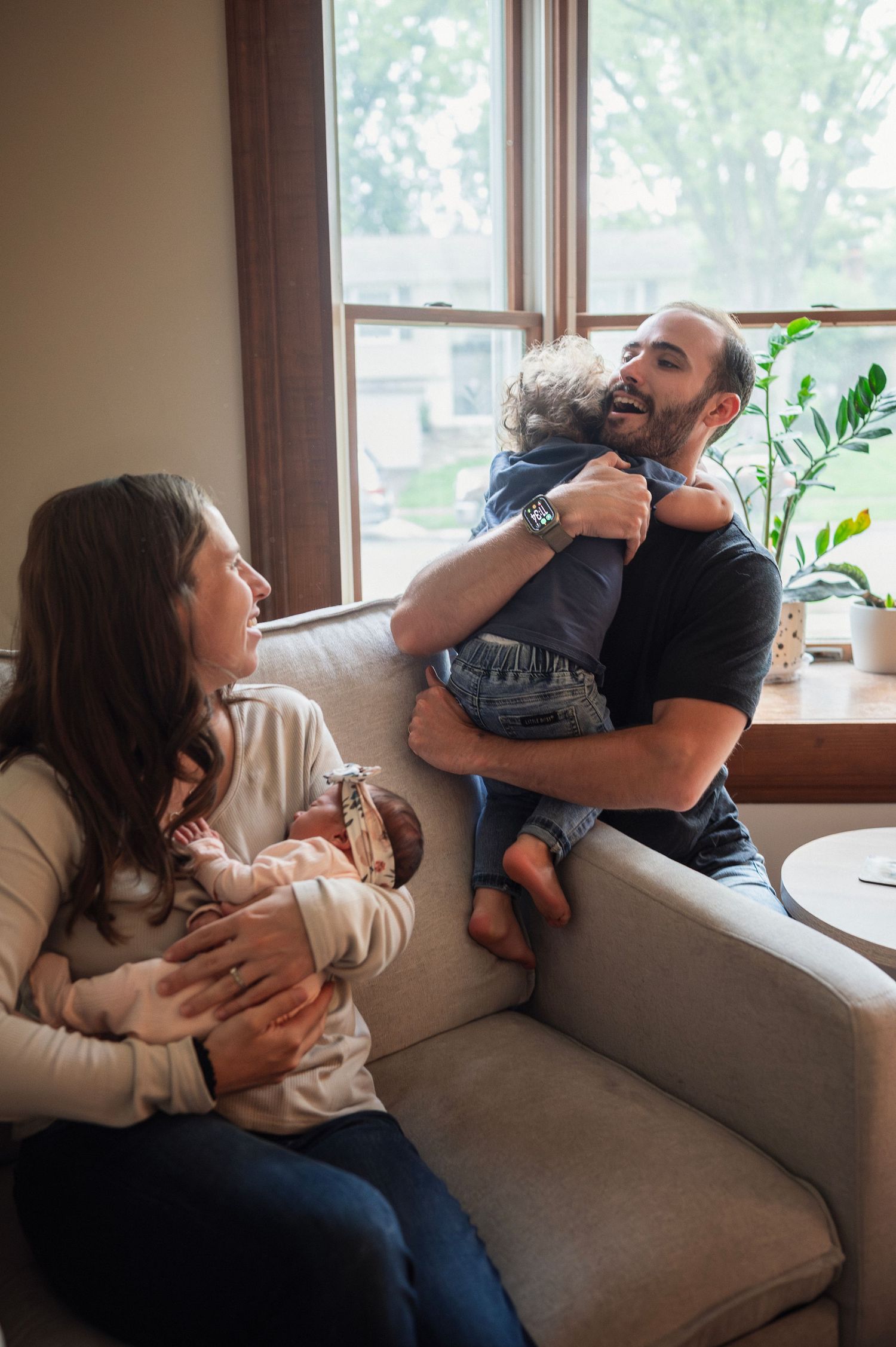 In-home Westerville session with dad hugging toddler and mom holding newborn in natural light.