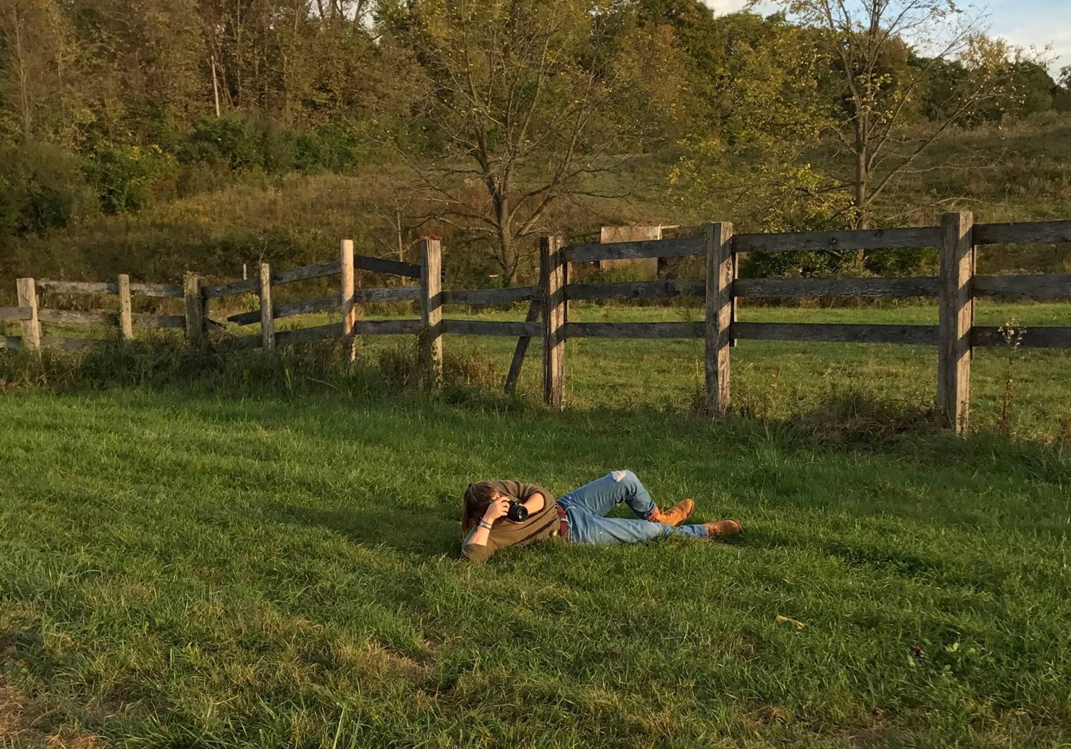 Photographer laying in the grass at golden hour with camera in hand, capturing the perfect low-angle shot beside a rustic wooden fence.
