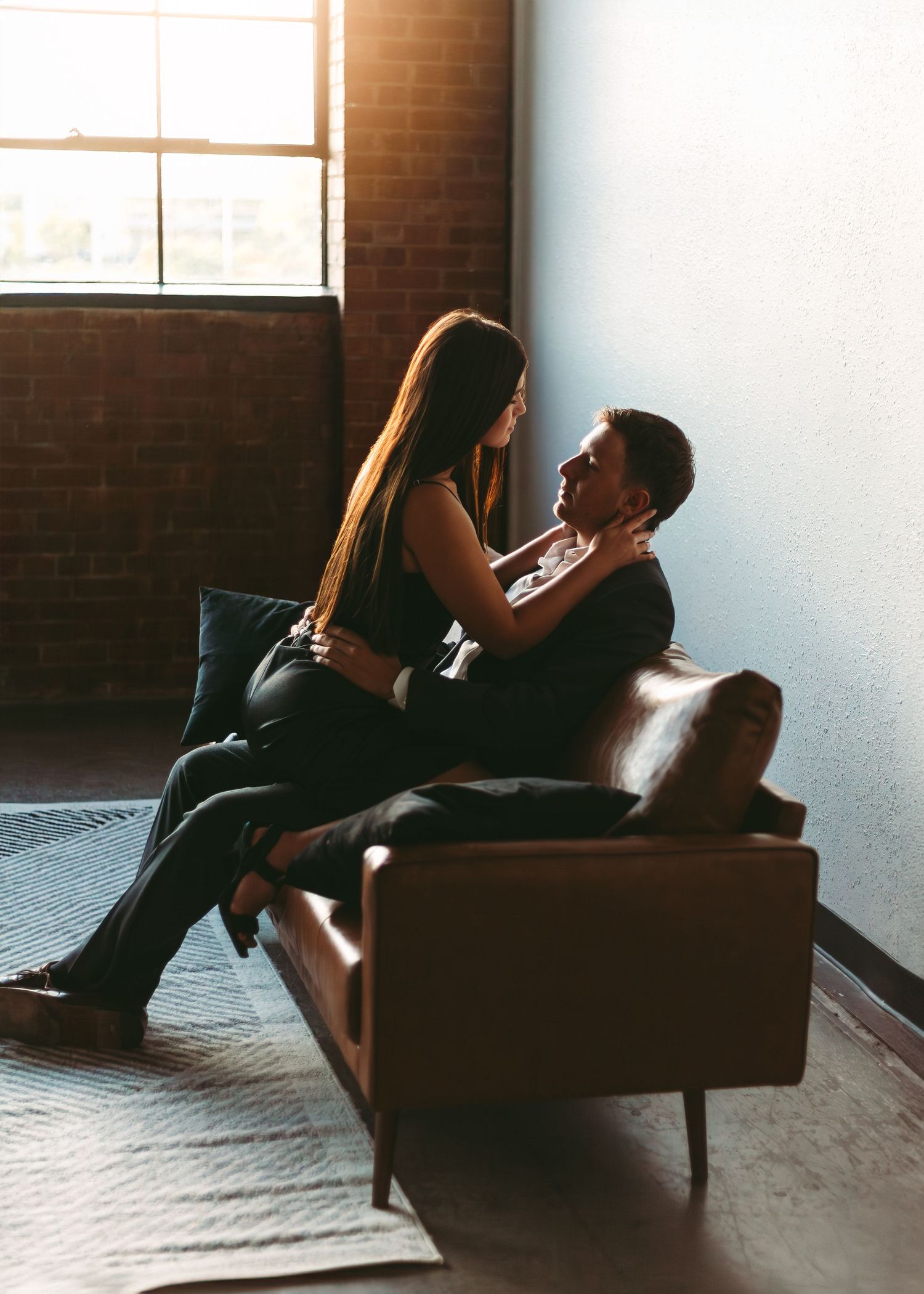 Romantic couple sitting intimately together on a brown leather sofa near a window with sunlight streaming in.