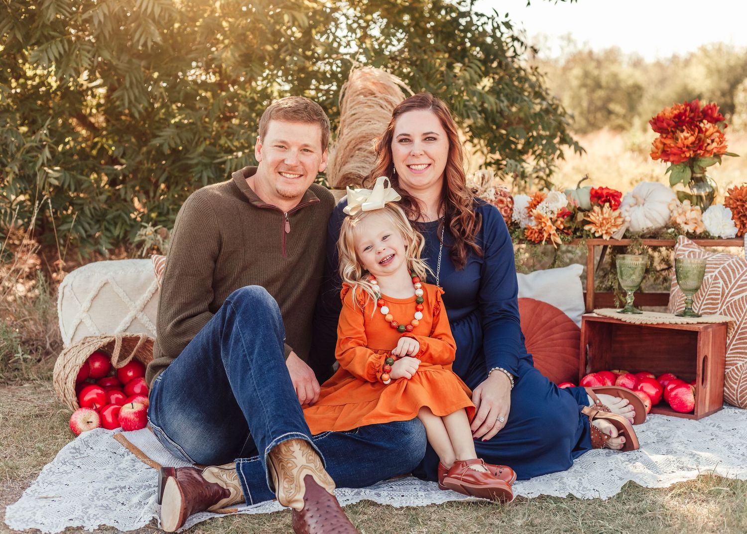 A family sits on a blanket outdoors during an autumn photoshoot with red decor and flowers in the background.