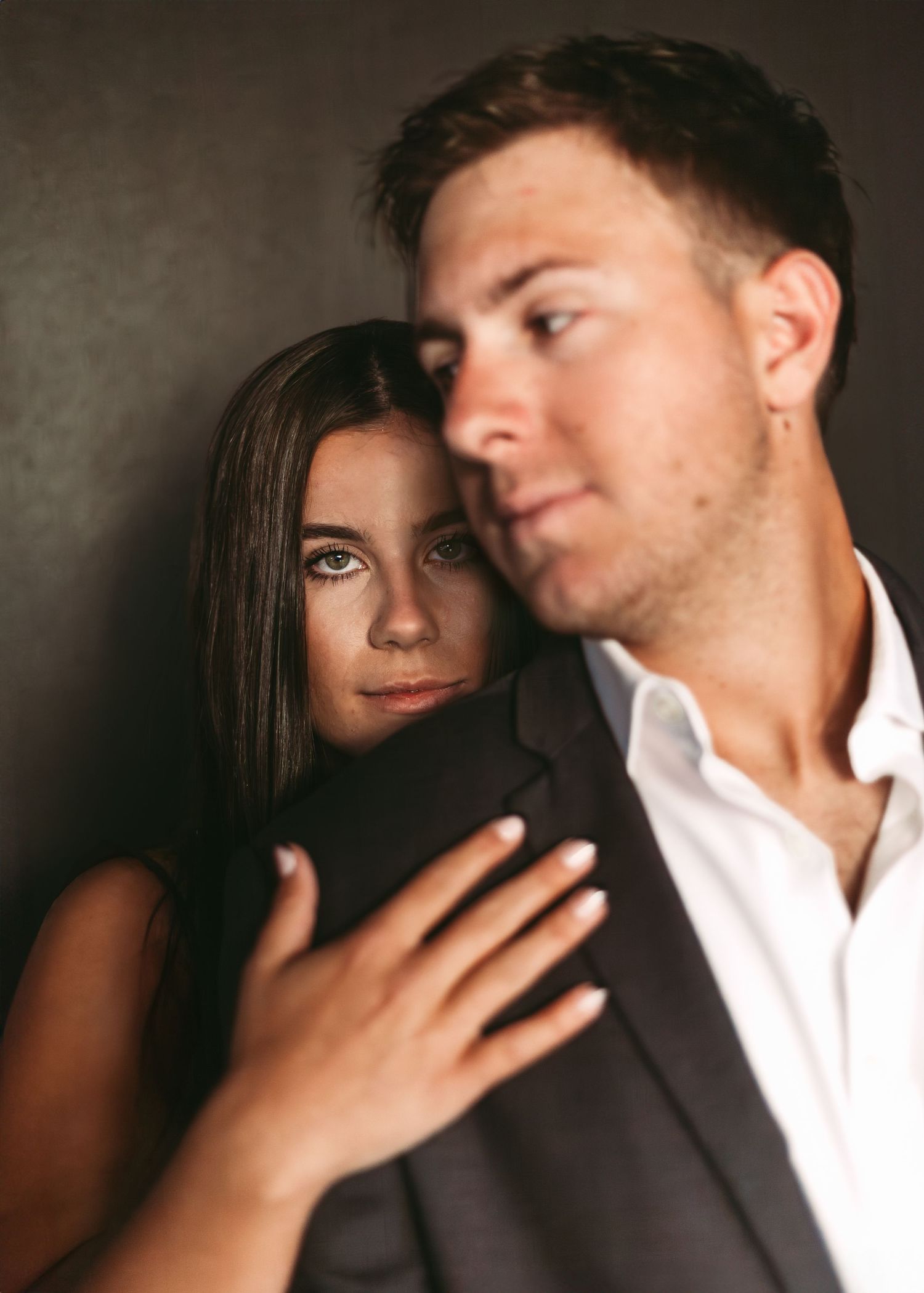Intimate portrait of a couple embracing in dramatic lighting against dark background, wearing formal attire.