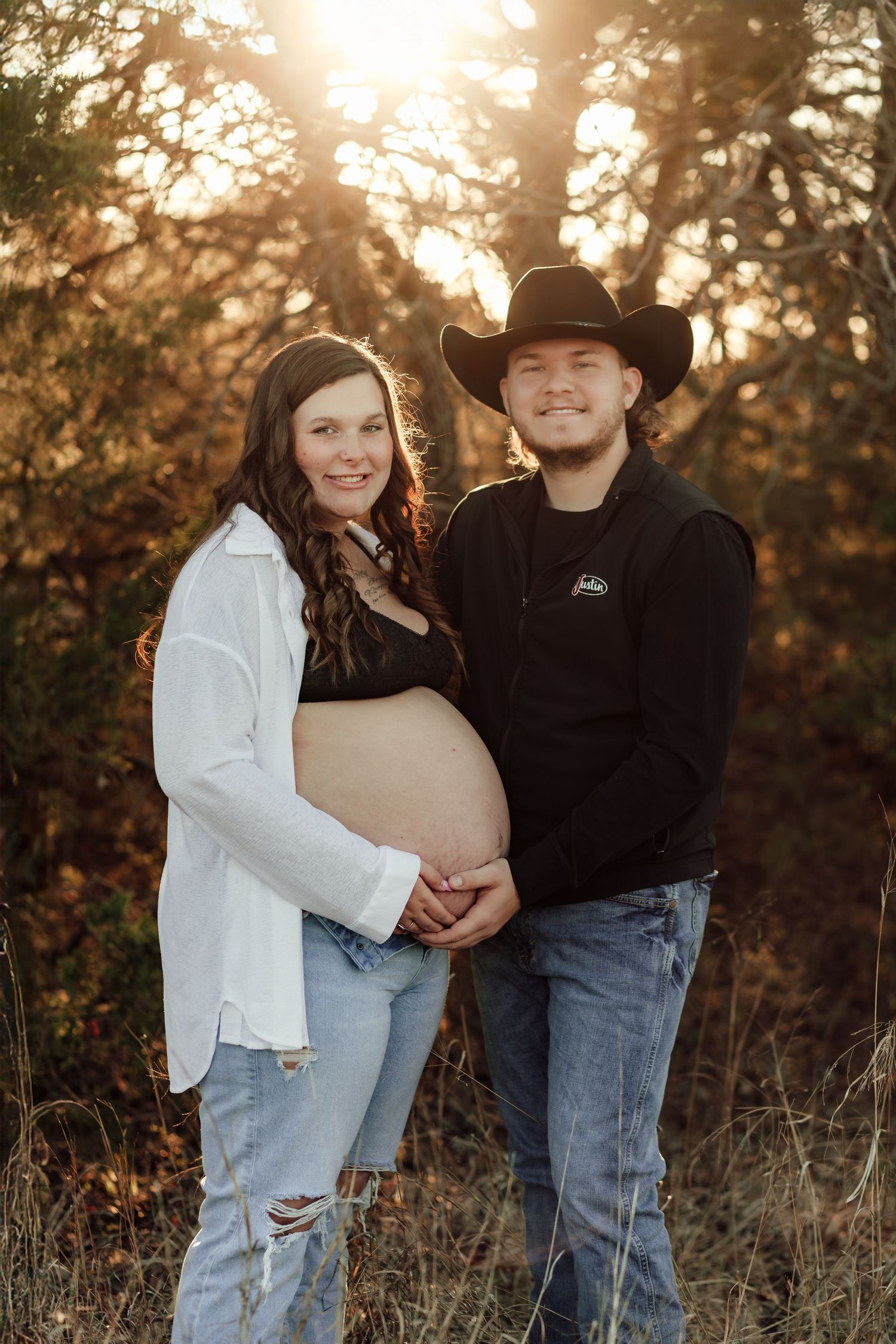 Expectant couple poses together outdoors at sunset, with a cowboy hat and autumn foliage in the background.