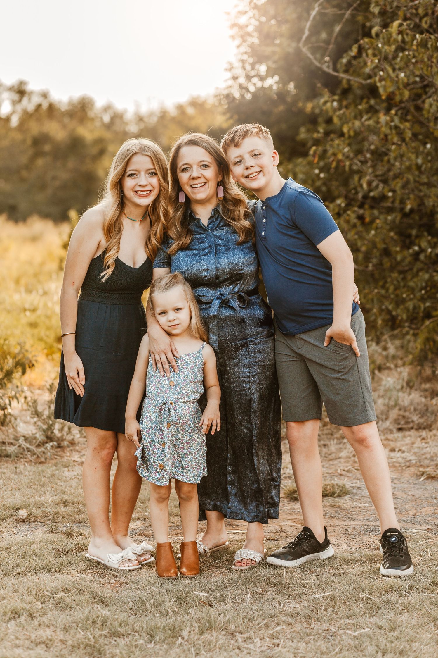 A family of four poses together outdoors in casual attire during golden hour sunset in a natural setting.