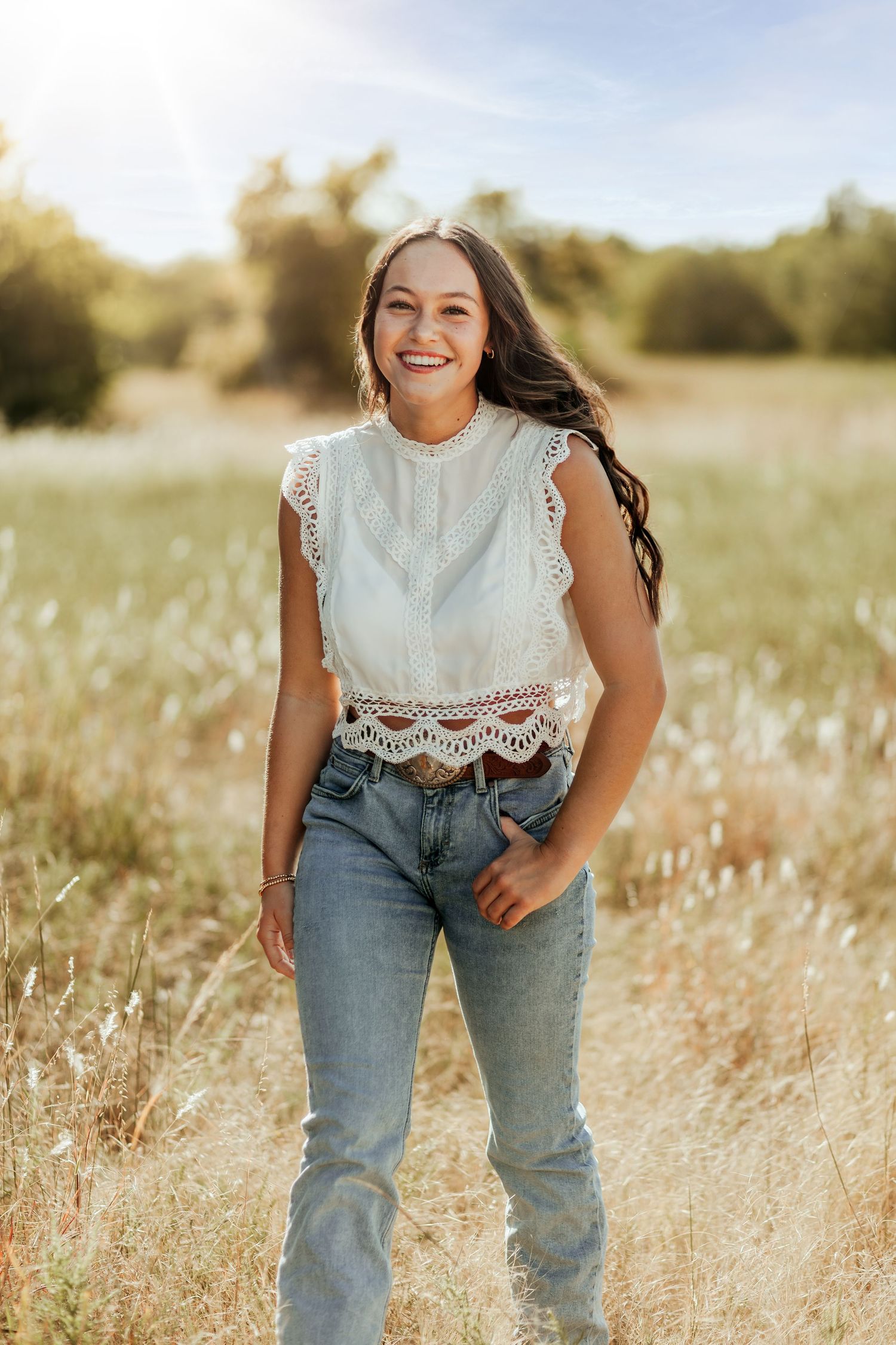 Woman in white lace top and blue jeans poses playfully in a sunlit field of tall golden grass.