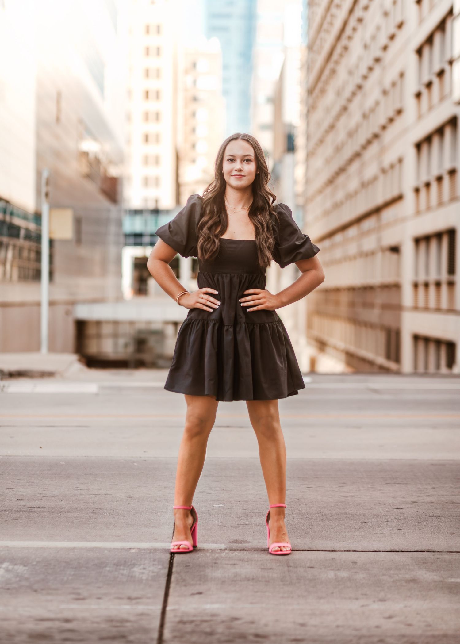 Someone in a black dress poses confidently on an urban sidewalk with tall buildings in the background at sunset.