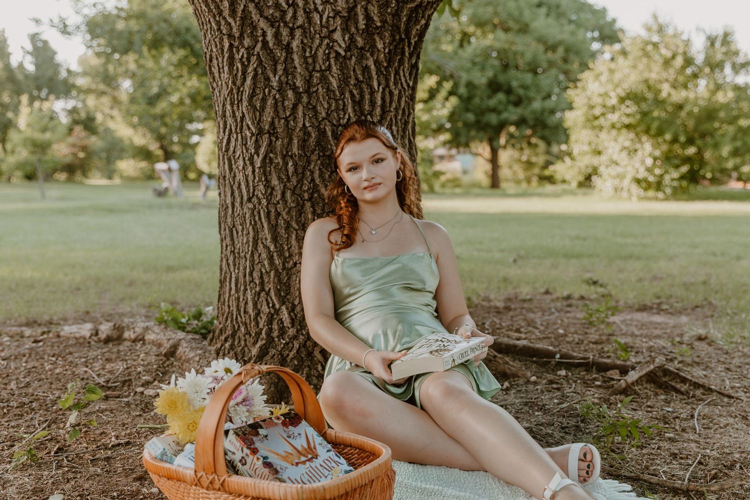 Someone in a mint green dress sits under a tree on a picnic blanket with a woven basket and flowers nearby.