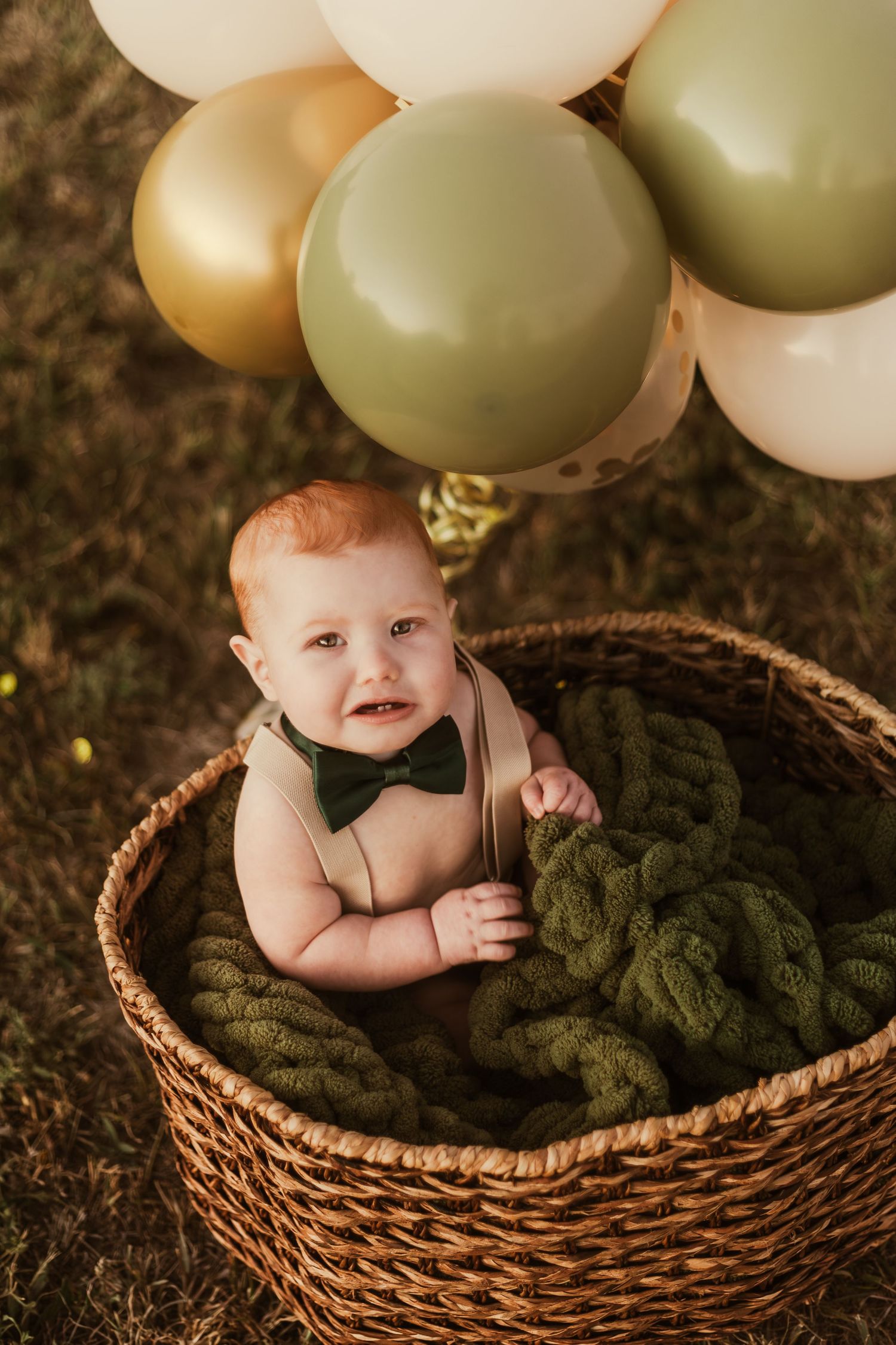 Smiling baby wearing bow tie sits in wicker basket with green blanket under sage and cream colored balloons.