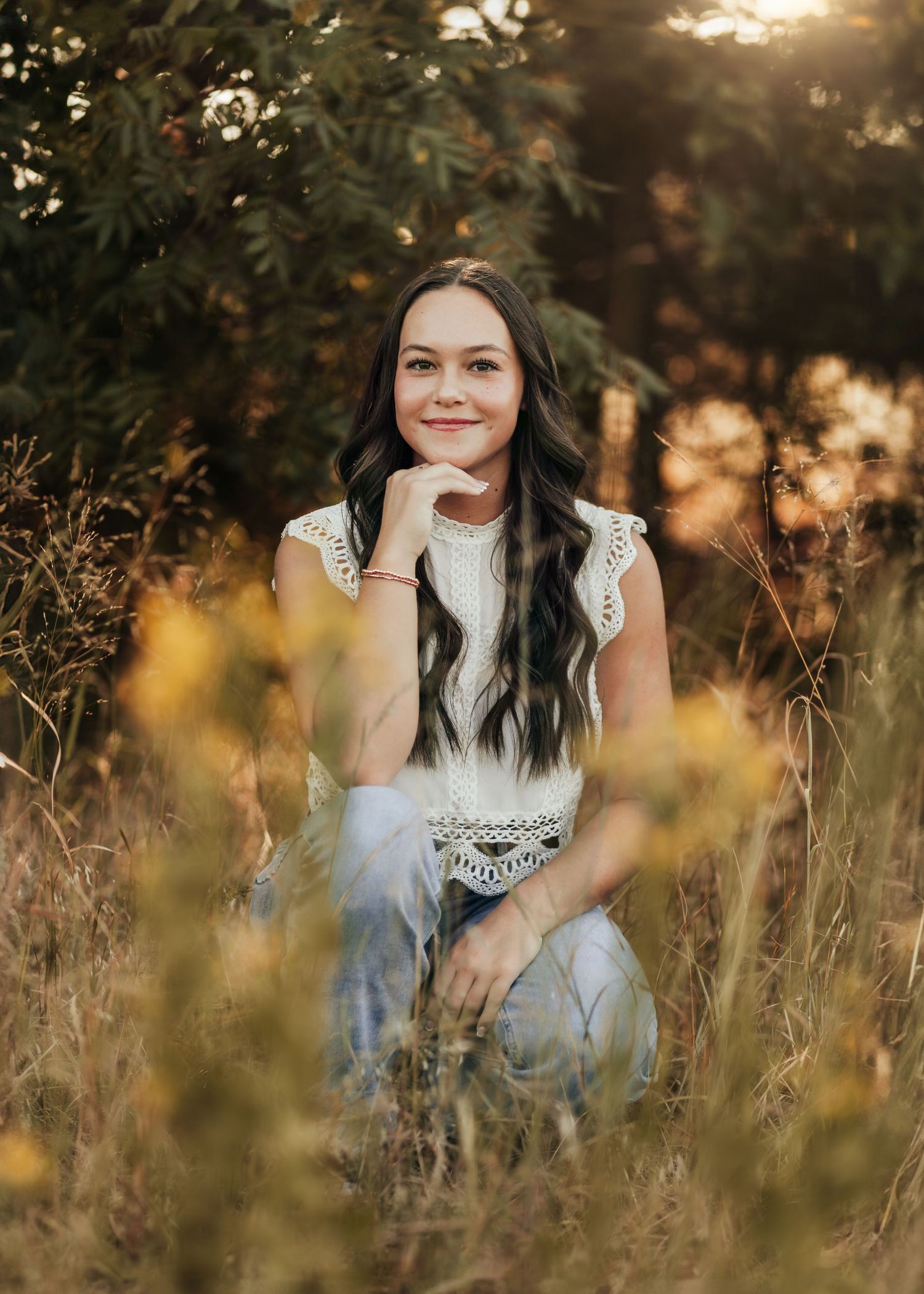 A woman in a white lace dress sits in golden sunlit field of tall grass during sunset.