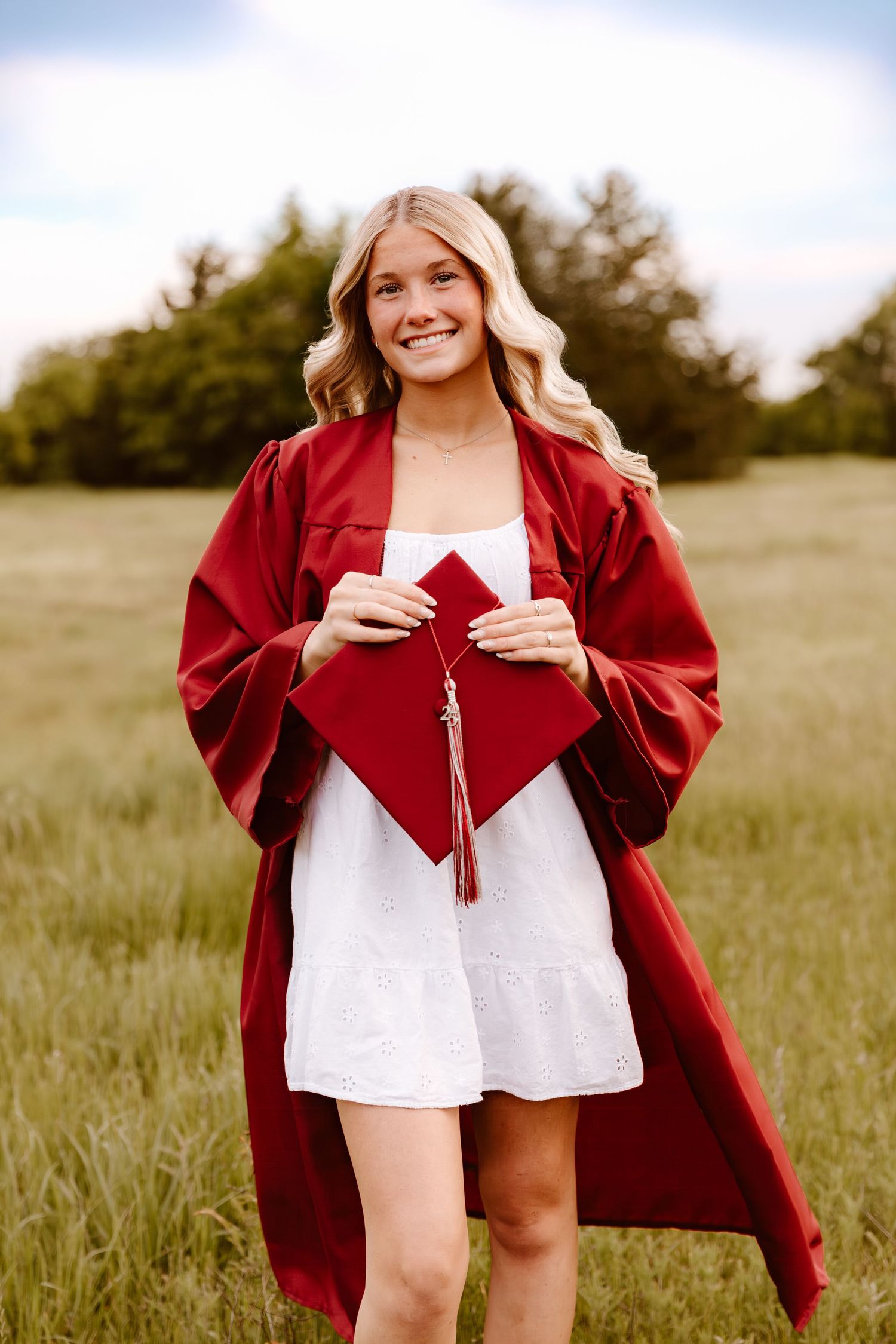 Graduate in red graduation gown holds cap and tassel while posing in grassy field during sunset.