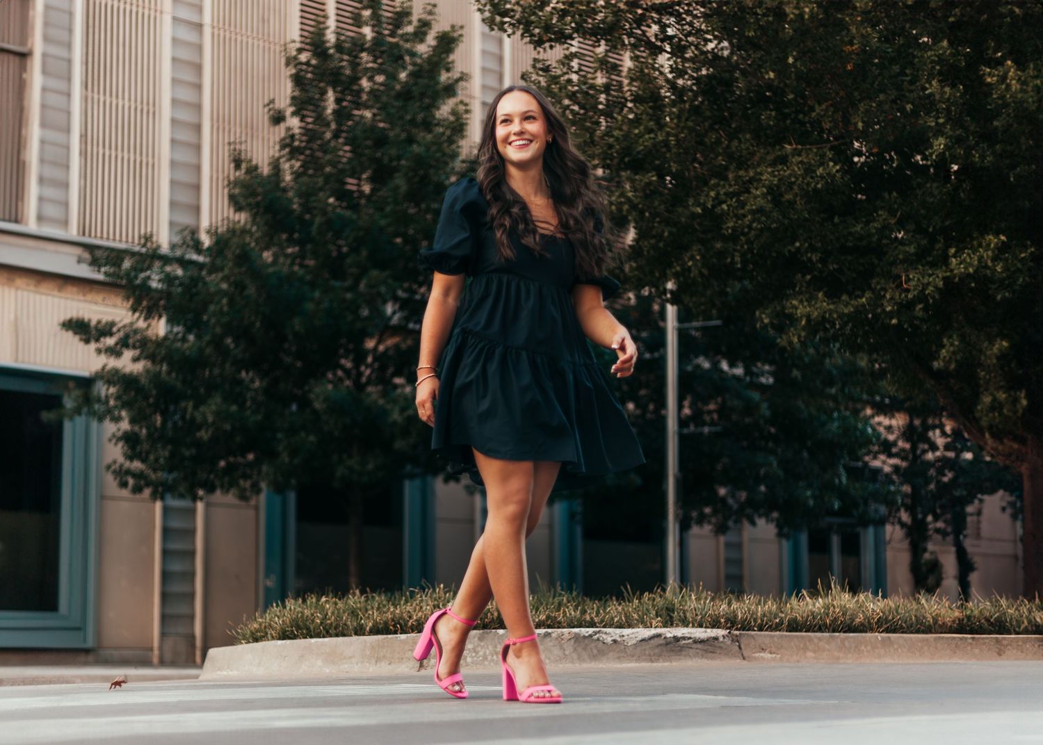 A person in a navy dress walks confidently down a city sidewalk wearing bright pink high heels.
