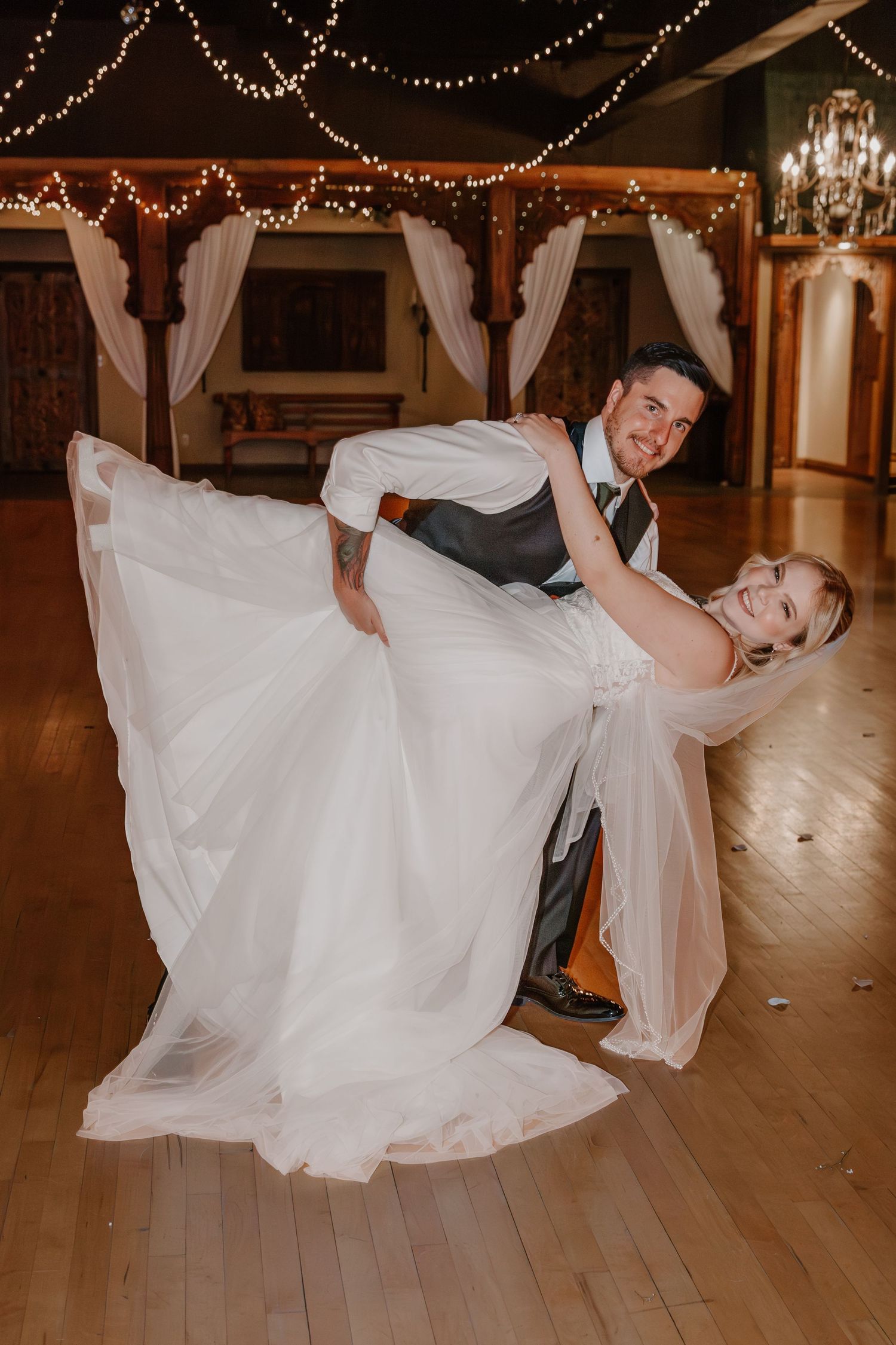 A couple dances during their wedding reception in a ballroom decorated with string lights and white drapes.