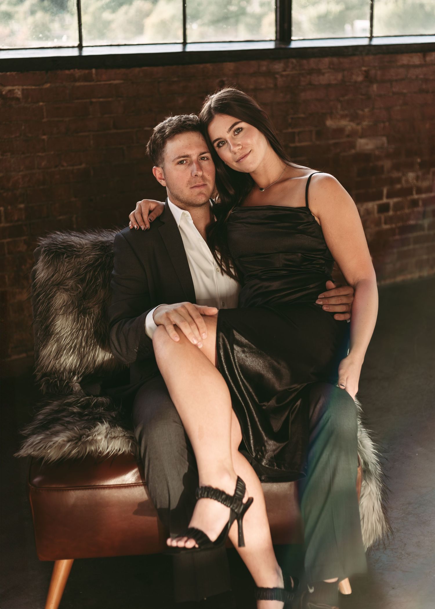 A romantic portrait of a couple in formal attire posing intimately on a leather couch against a brick wall backdrop.