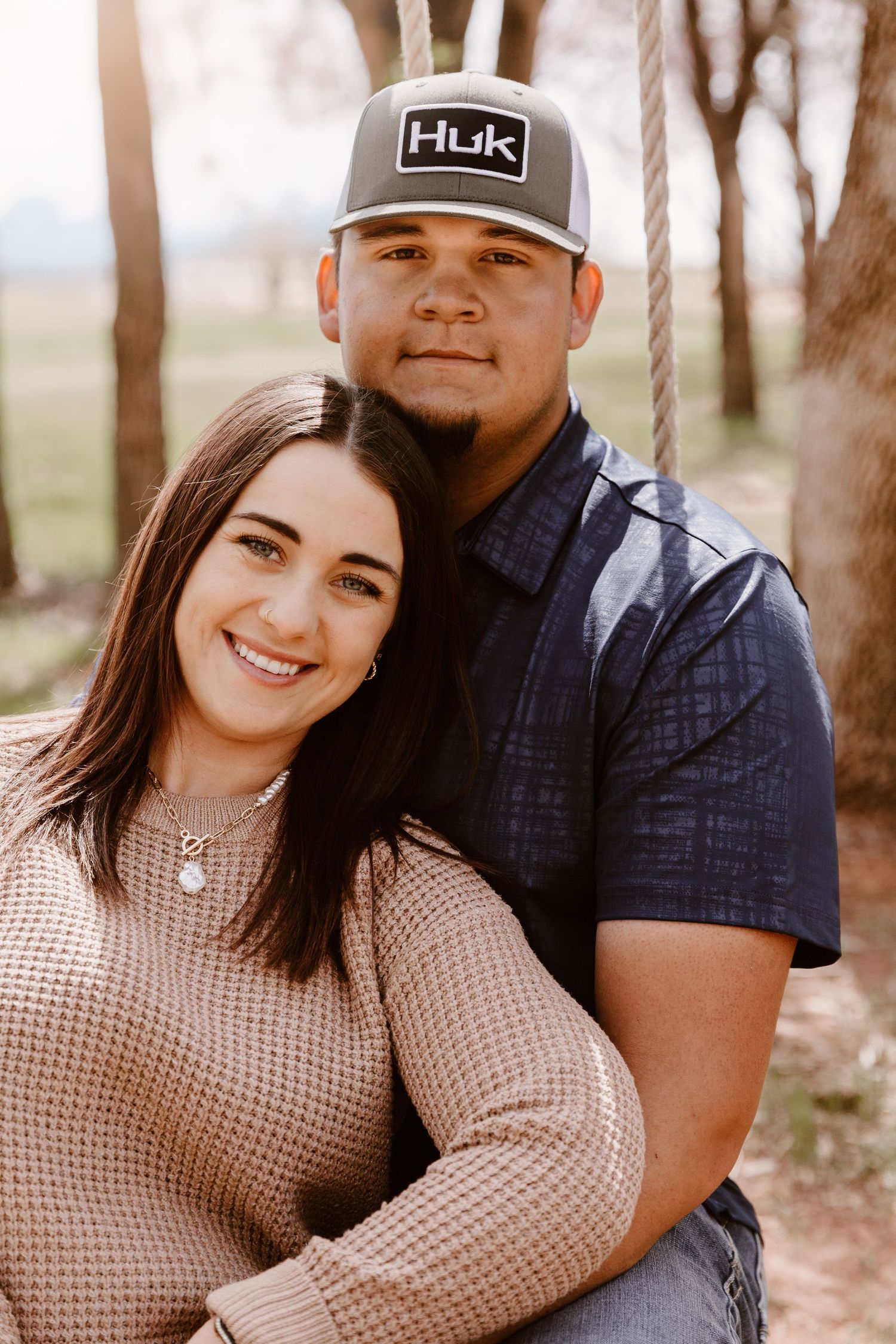 A couple poses outdoors in casual wear, with her in a beige sweater and him wearing a Huk baseball cap.