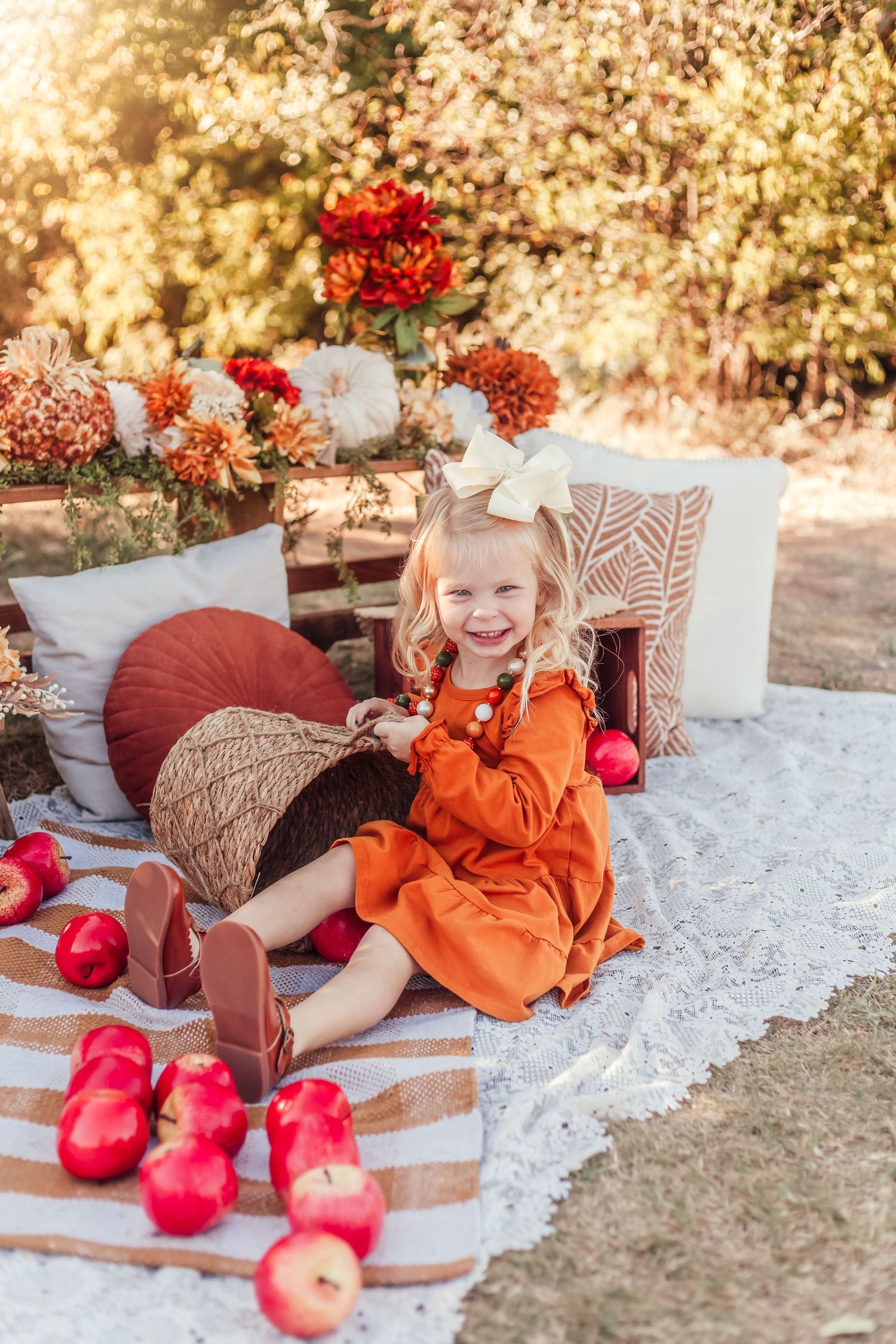 Cozy fall picnic setup with orange dress, red apples, throw pillows and floral arrangements in warm autumn sunlight.