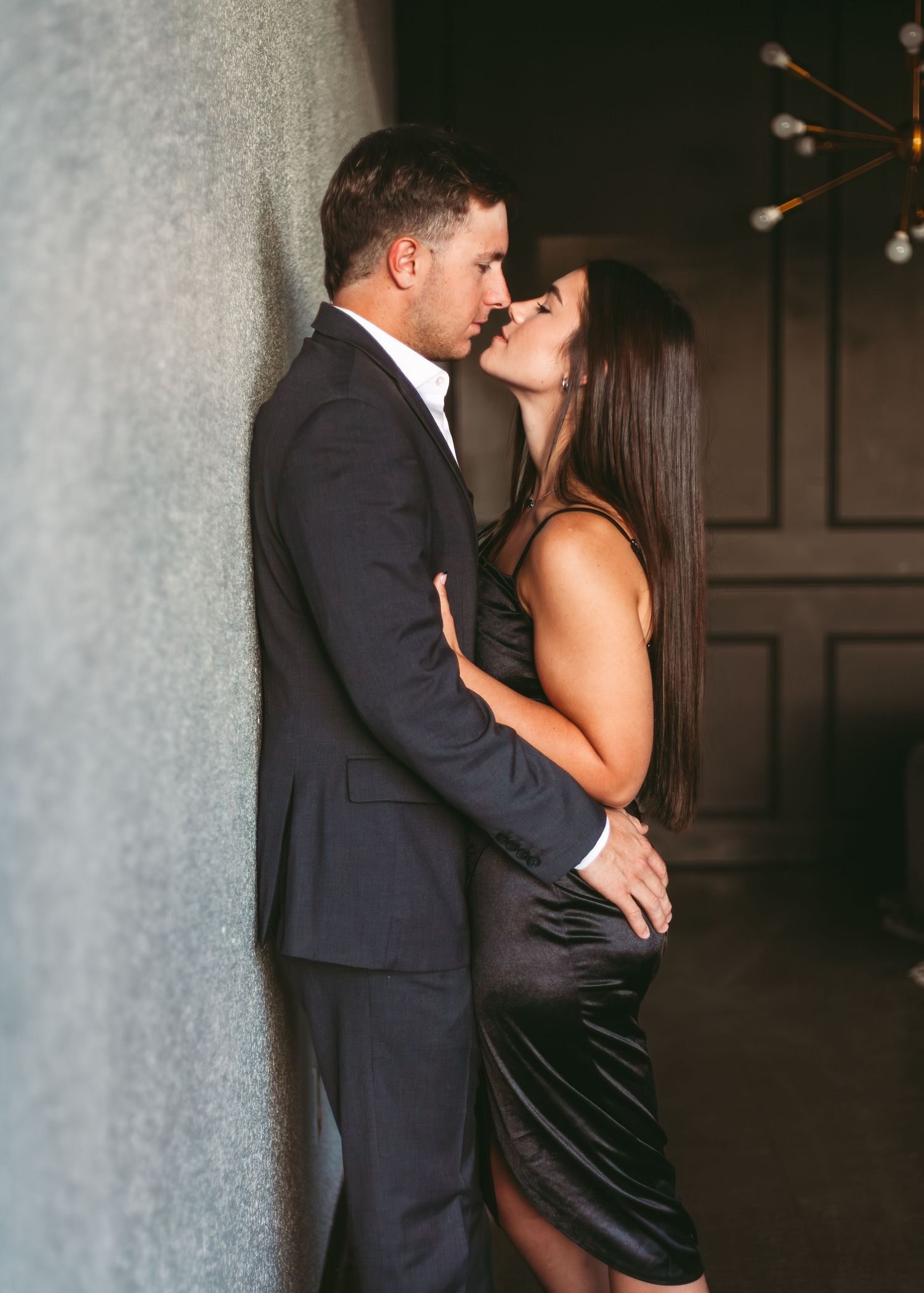 A couple in formal attire shares an intimate moment against a dark wall with a modern chandelier in the background.