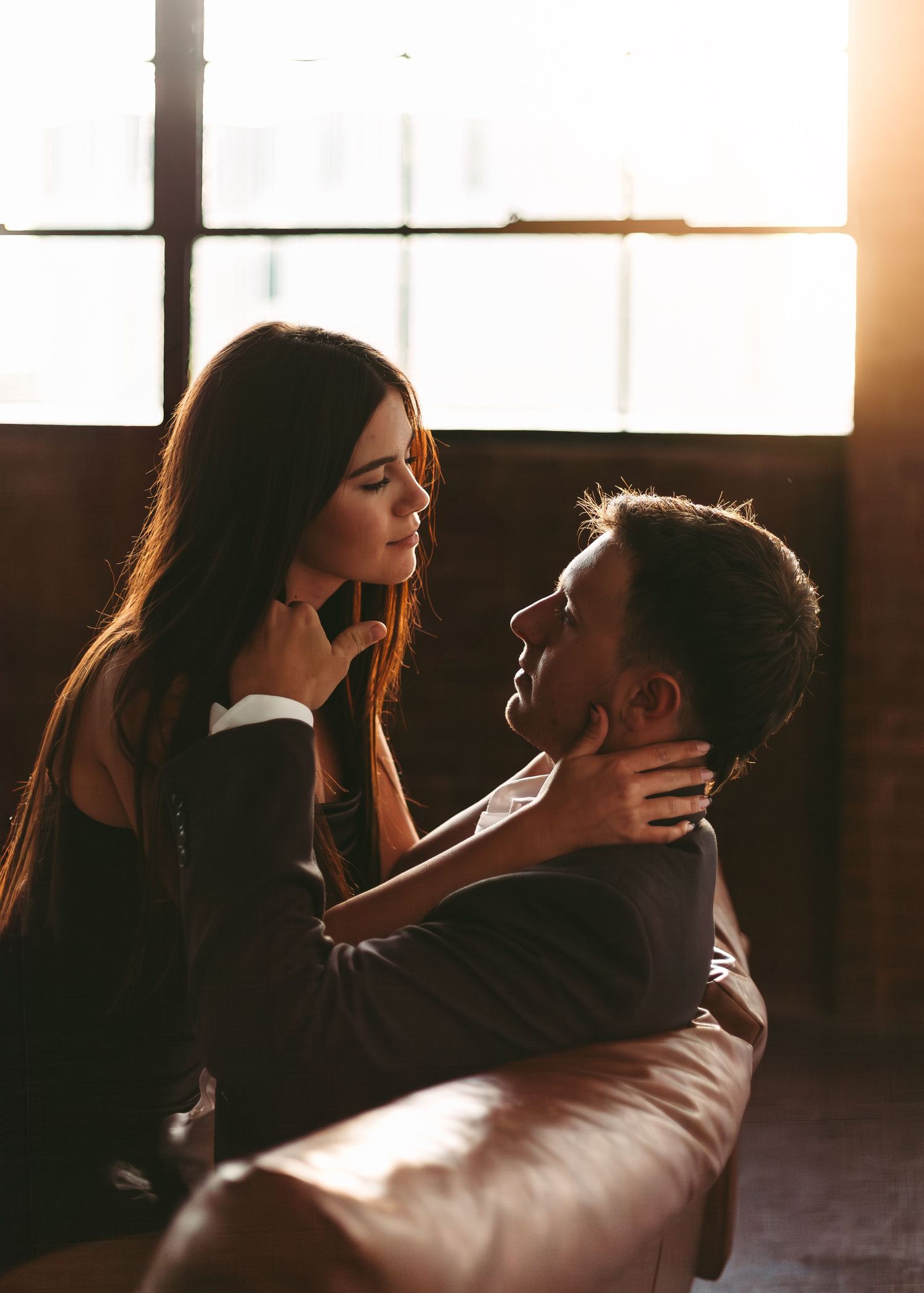 A romantic couple shares an intimate moment in warm lighting by a window, their silhouettes backlit in golden sunlight.